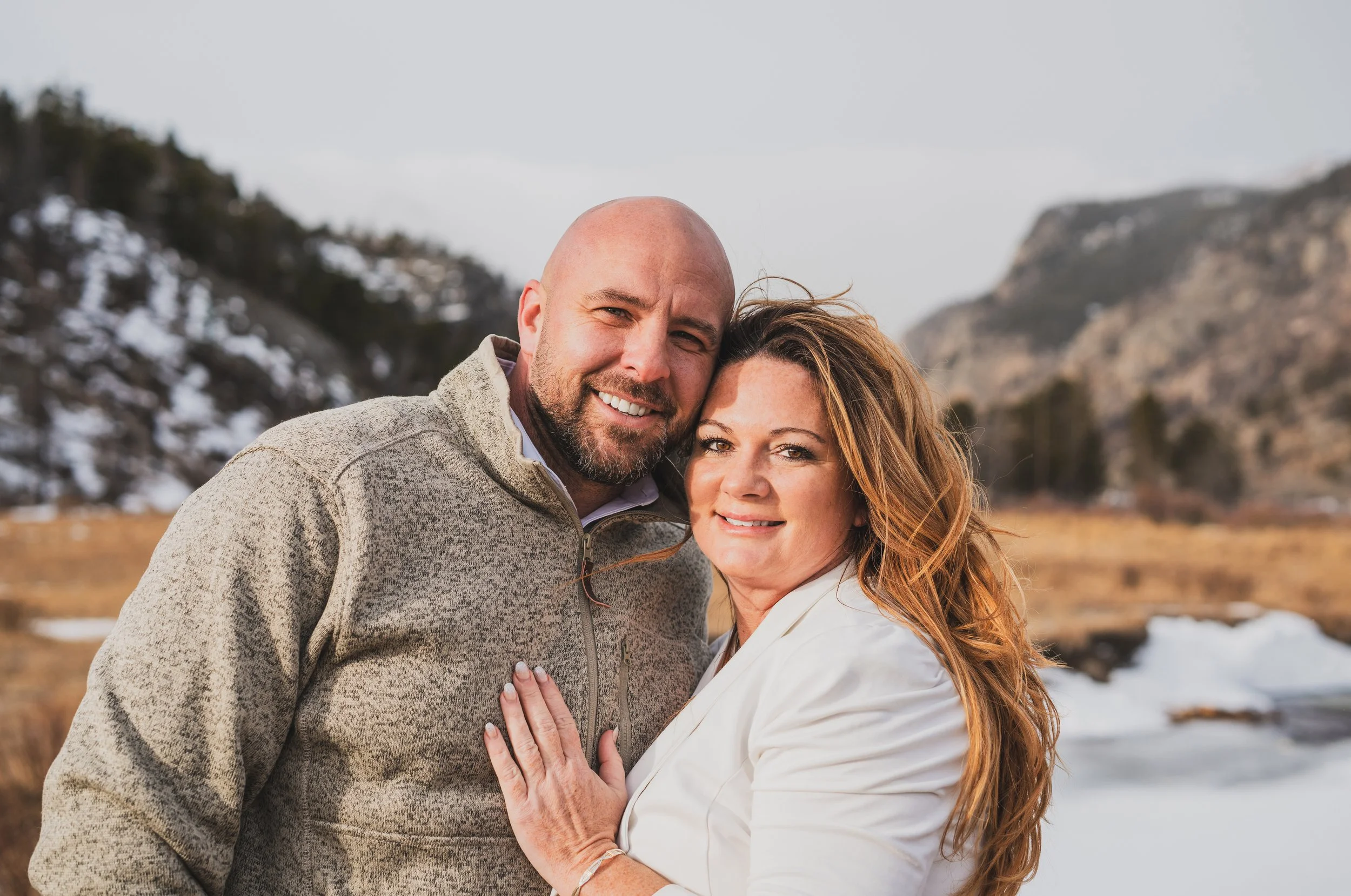 A smiling couple standing outdoors in a mountainous area with snow on the ground, embracing each other.