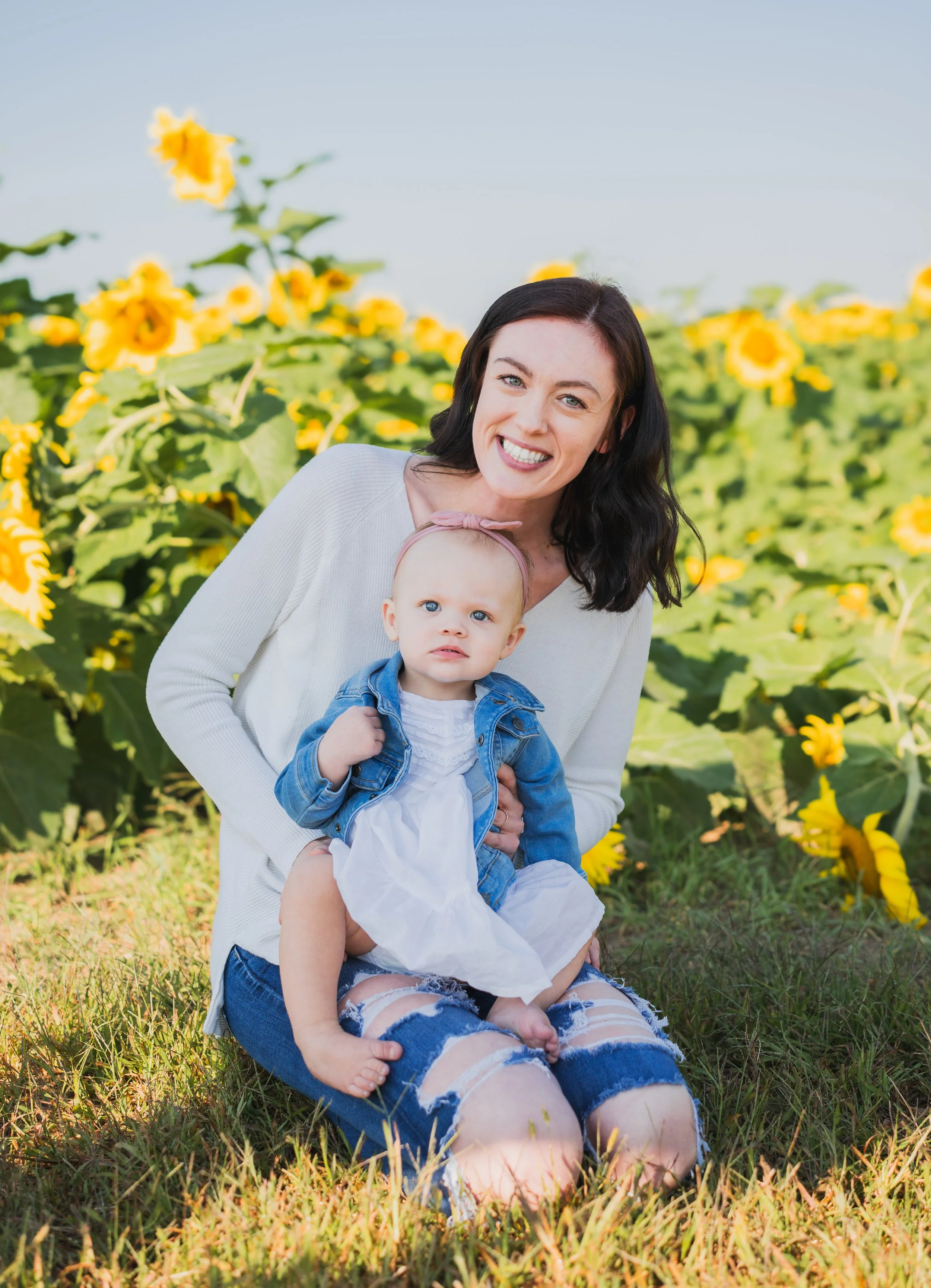 A woman and a young girl sitting on grass in a sunflower field smiling at the camera.