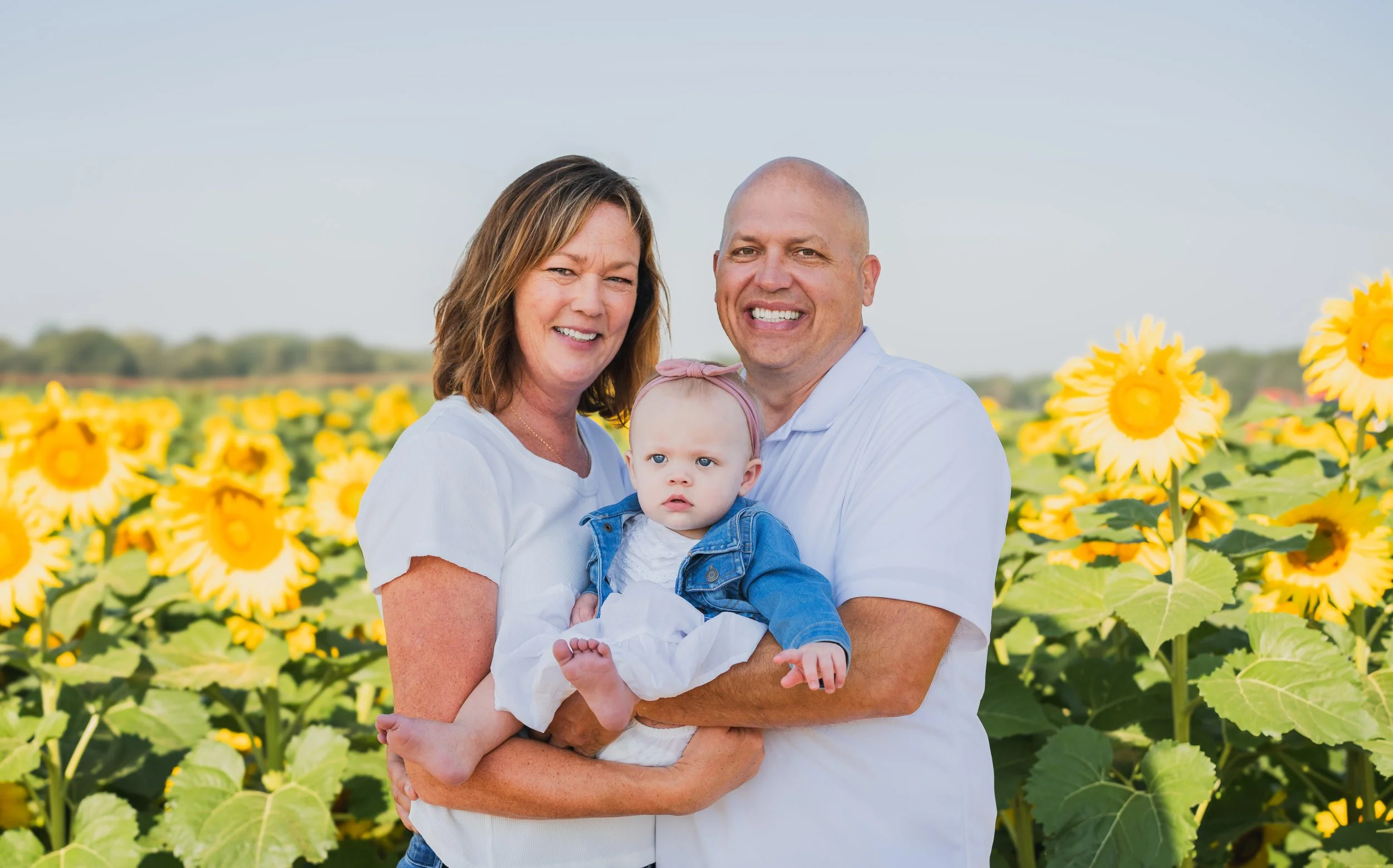 A family of three standing in a sunflower field, smiling at the camera. The woman has shoulder-length brown hair, the man is bald, and the baby girl has a pink headband and is wearing a denim jacket.