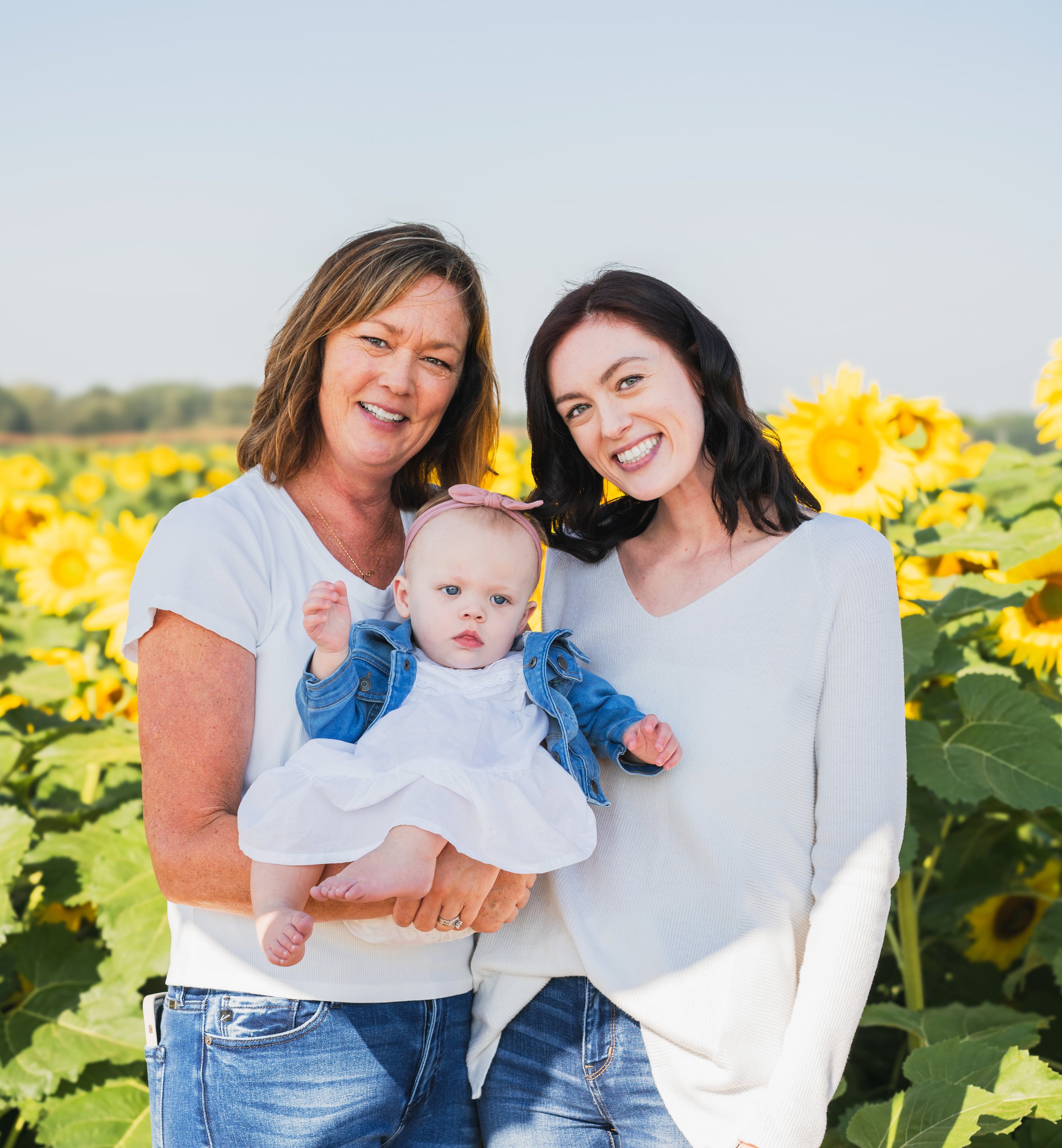 Three women, including a baby, standing in a sunflower field, smiling at the camera.
