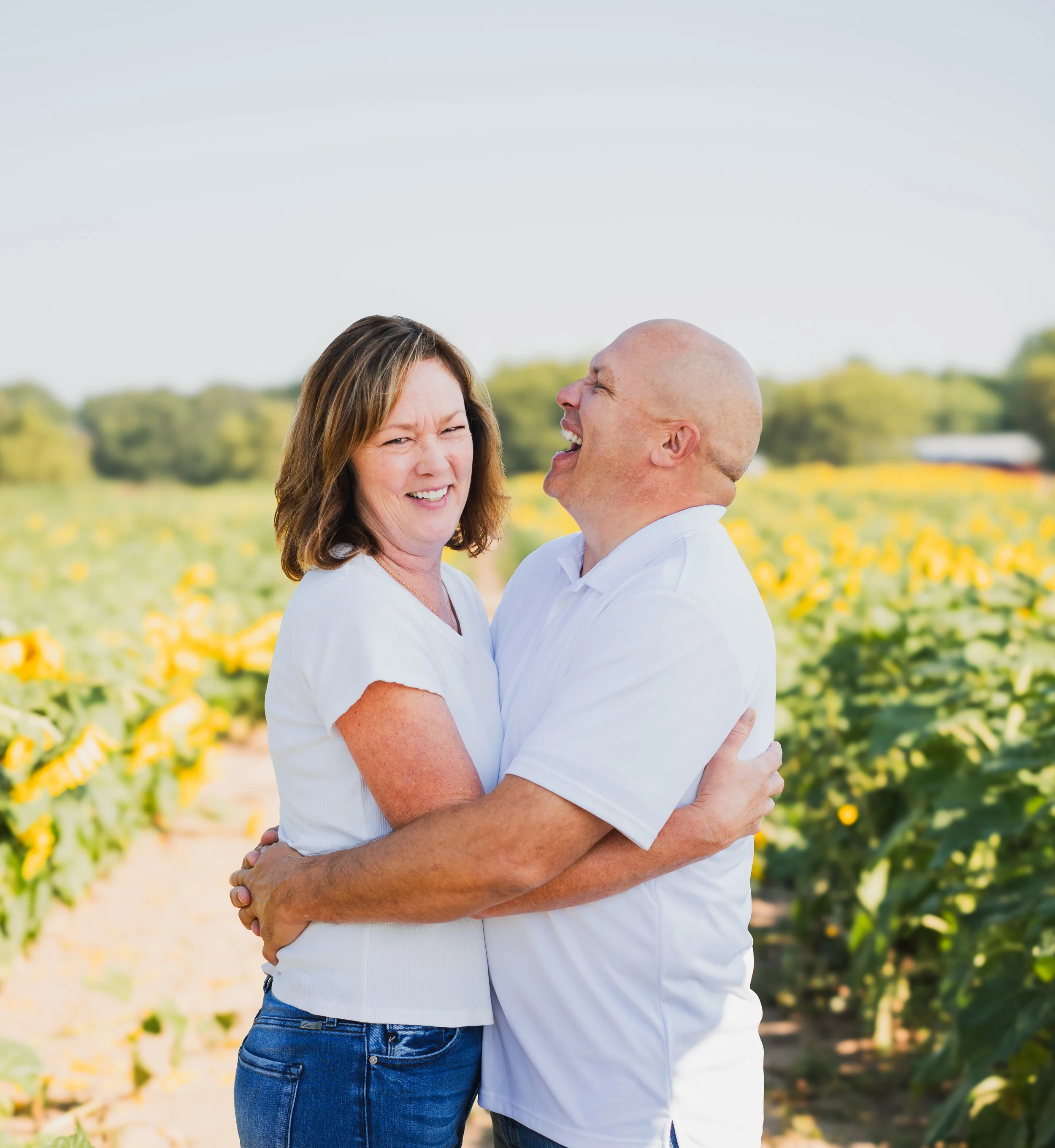 A couple laughing and hugging each other in a sunflower field on a sunny day.