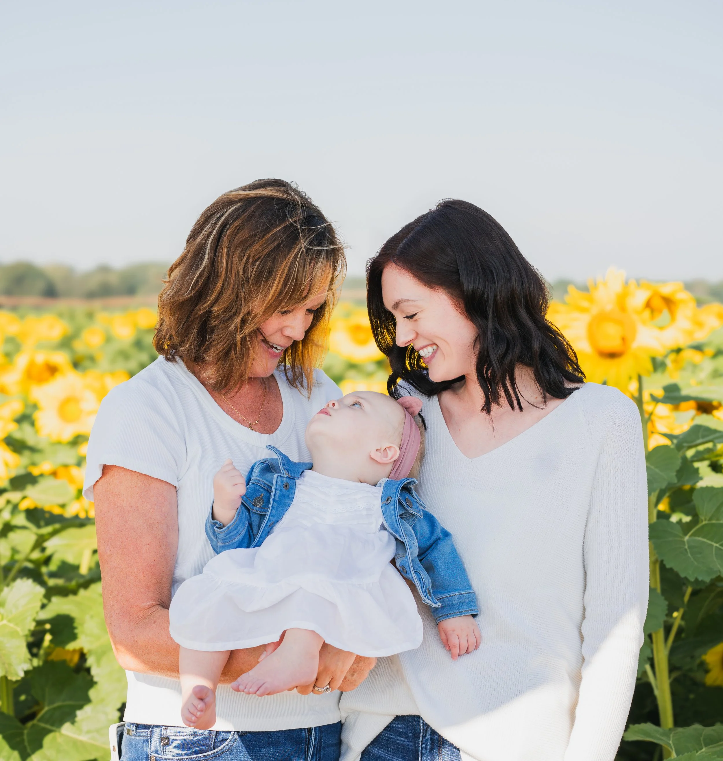 Two women holding a baby girl in a sunflower field, smiling at each other.