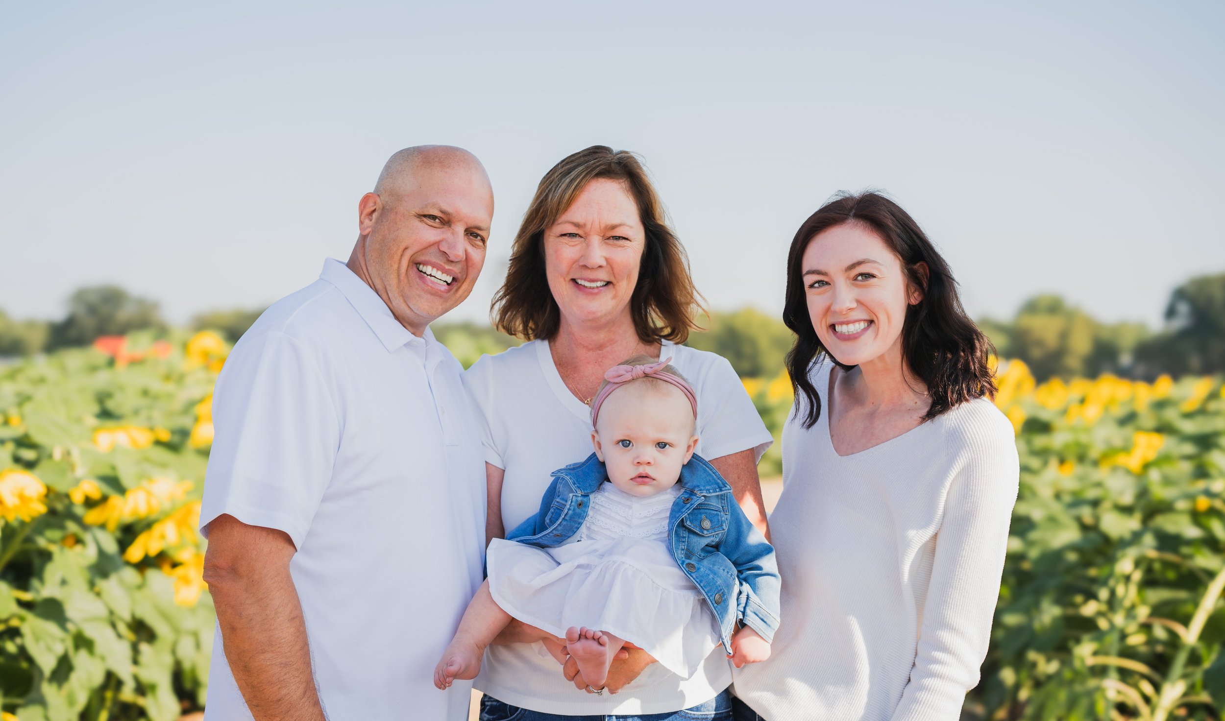 A happy multi-generational family standing outdoors in a field of sunflowers on a bright, sunny day, smiling at the camera.