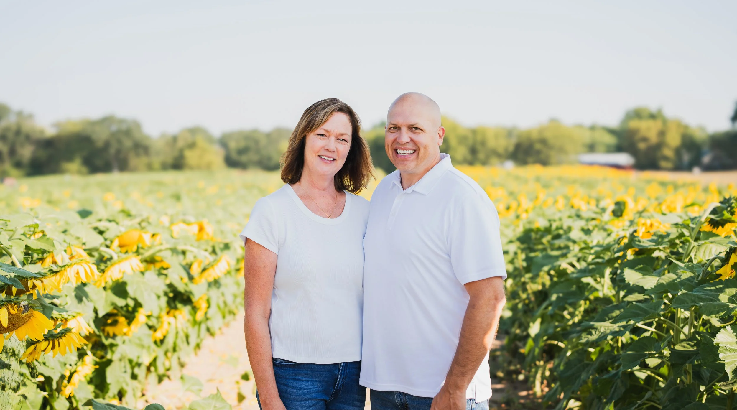 A smiling couple standing in a sunflower field on a sunny day with green trees in the background.