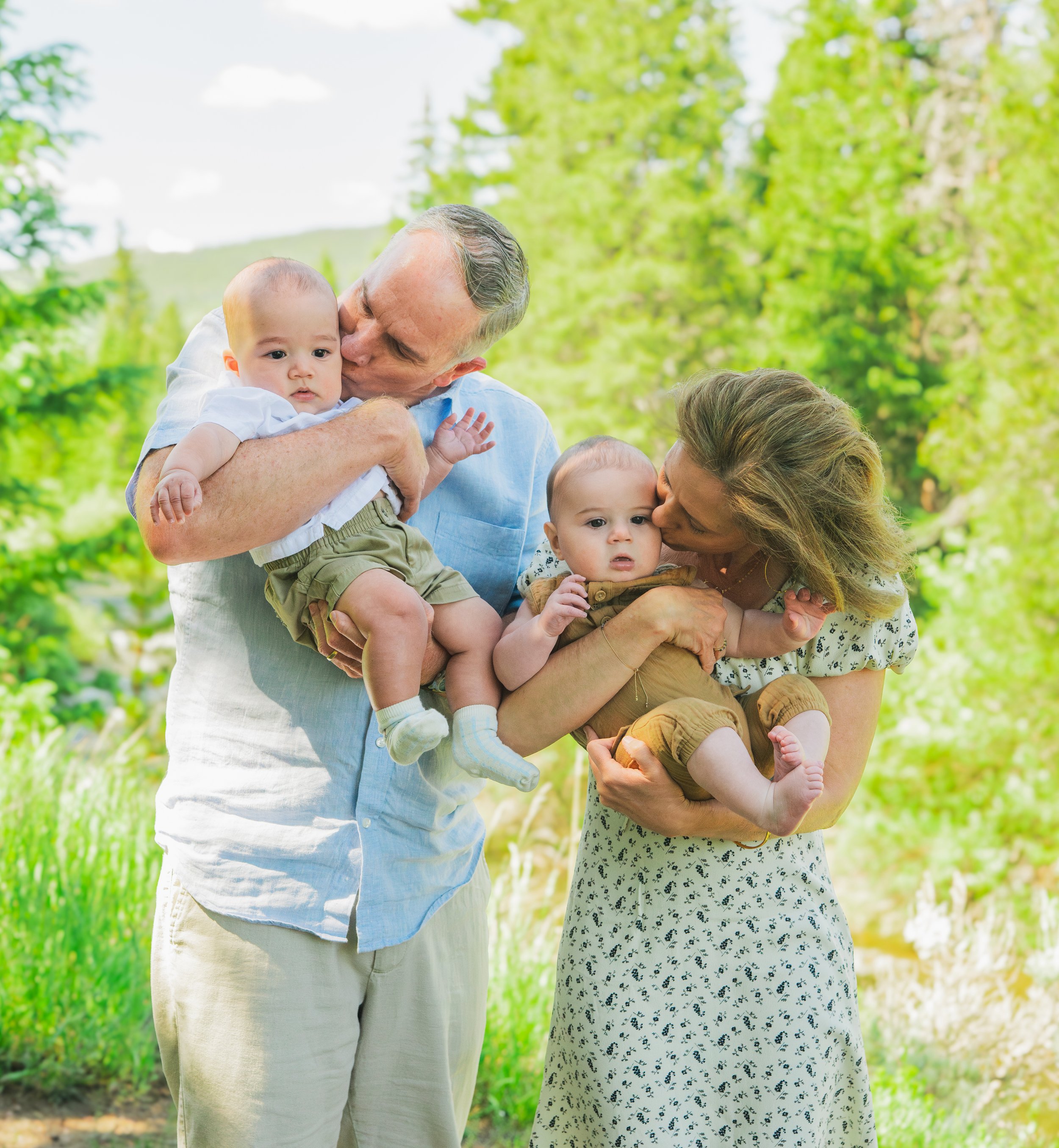 A family of four with grandparents and two young children outdoors in a green, wooded area, sharing kisses and holding the children.