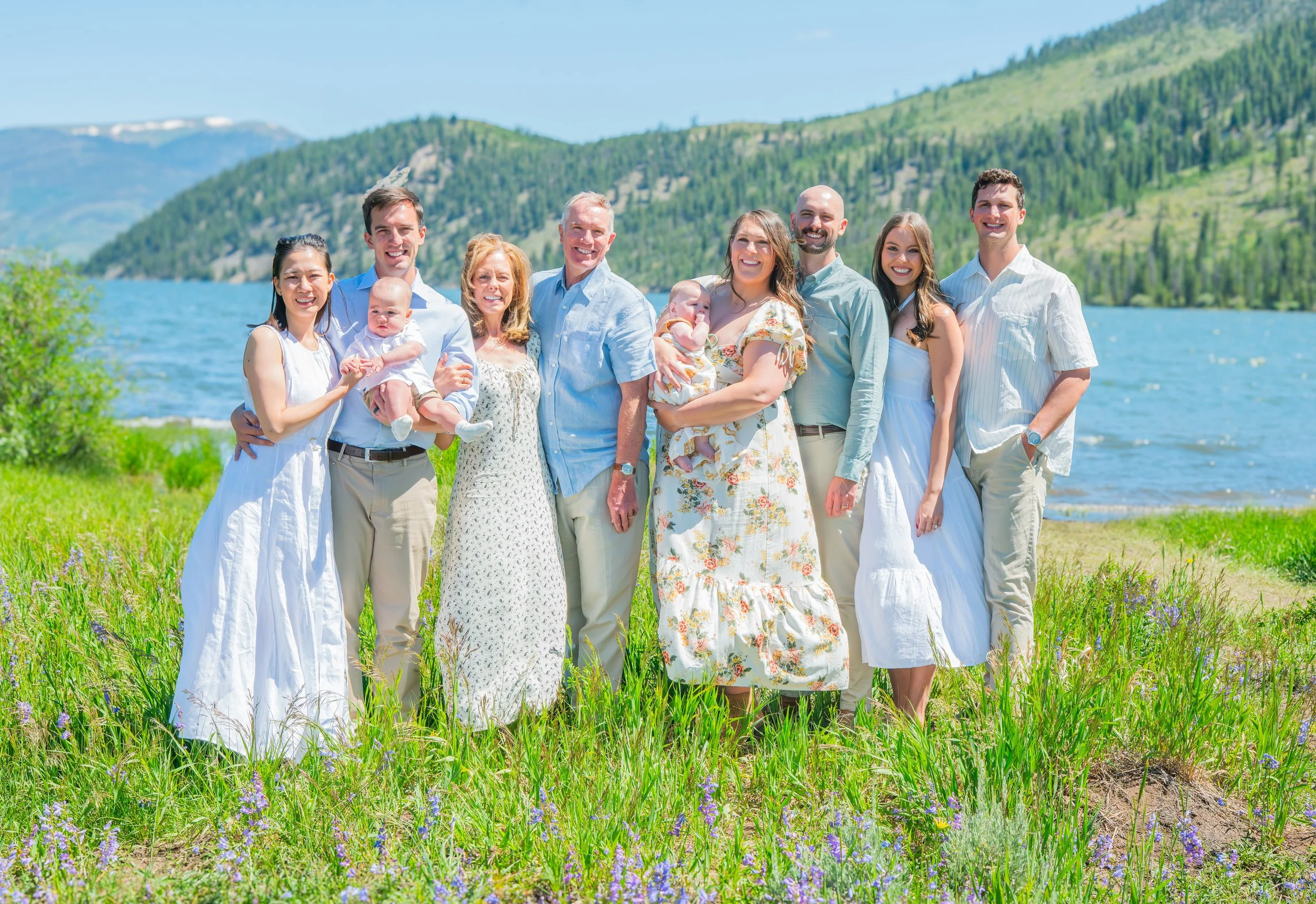 Group of ten people standing on grass near a lake with mountains in the background, smiling for a photo on a sunny day.