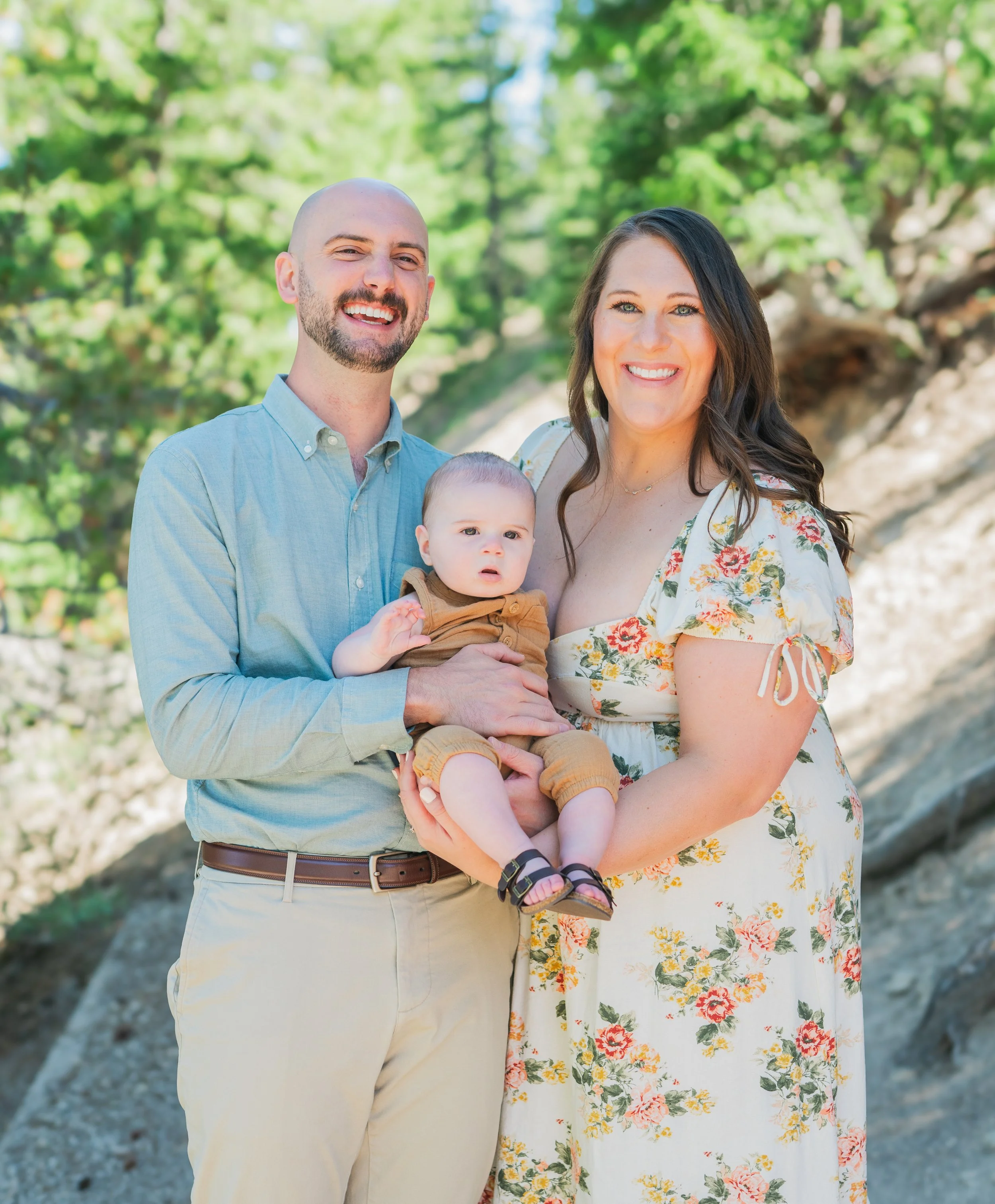 A happy family of three standing outdoors in a sunlit, green forest, with the man holding a young child and the woman smiling next to them.