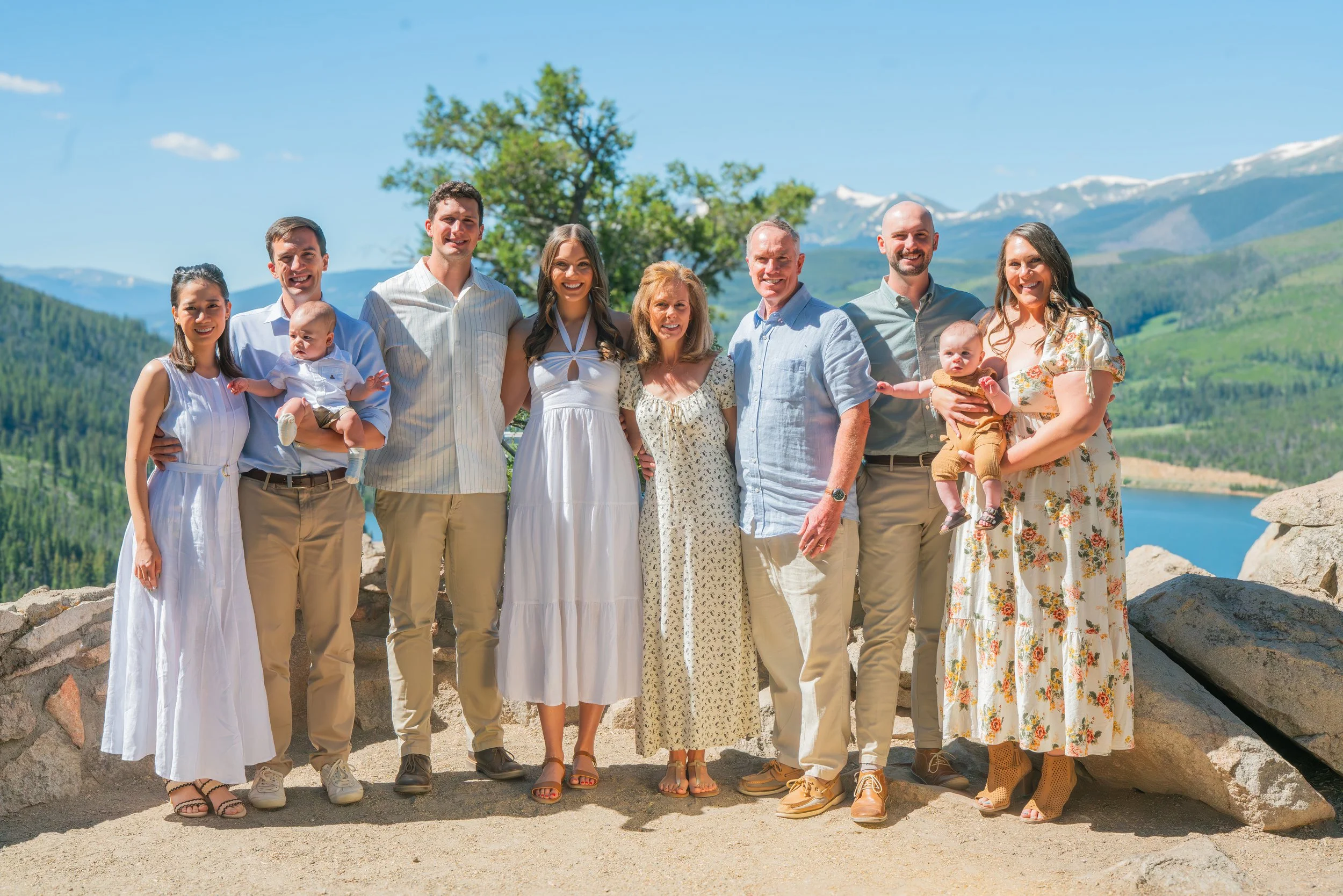 Group of ten people, including children, standing outdoors with mountains and a lake in the background on a sunny day.