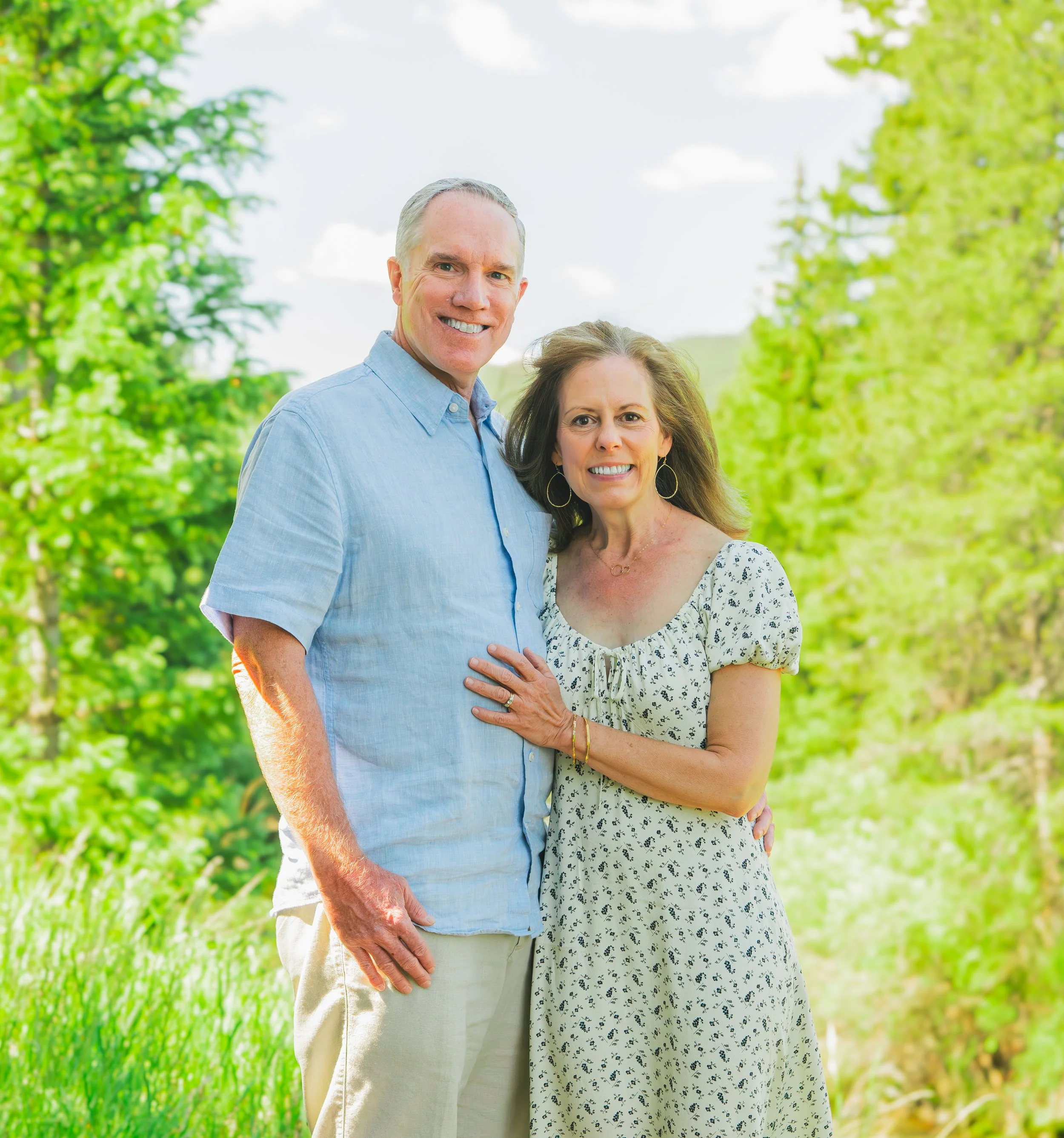 A smiling middle-aged couple standing outdoors in a green, leafy area on a sunny day.