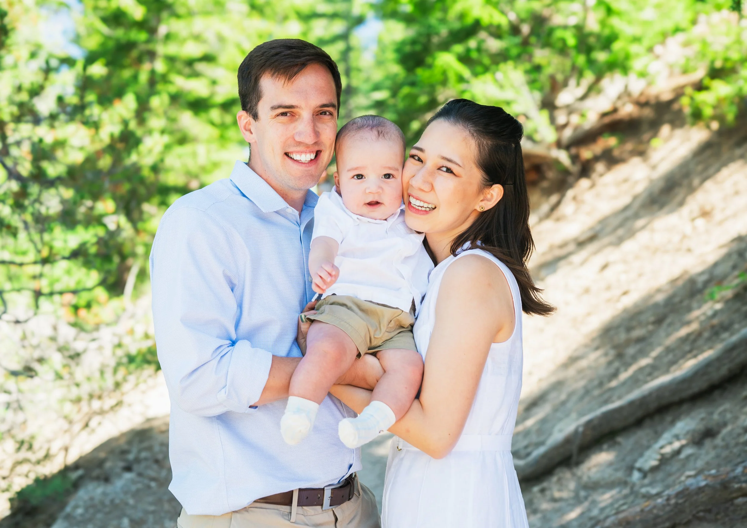 A happy family of three, including a father, mother, and baby, outdoors on a sunny day, smiling and holding the baby