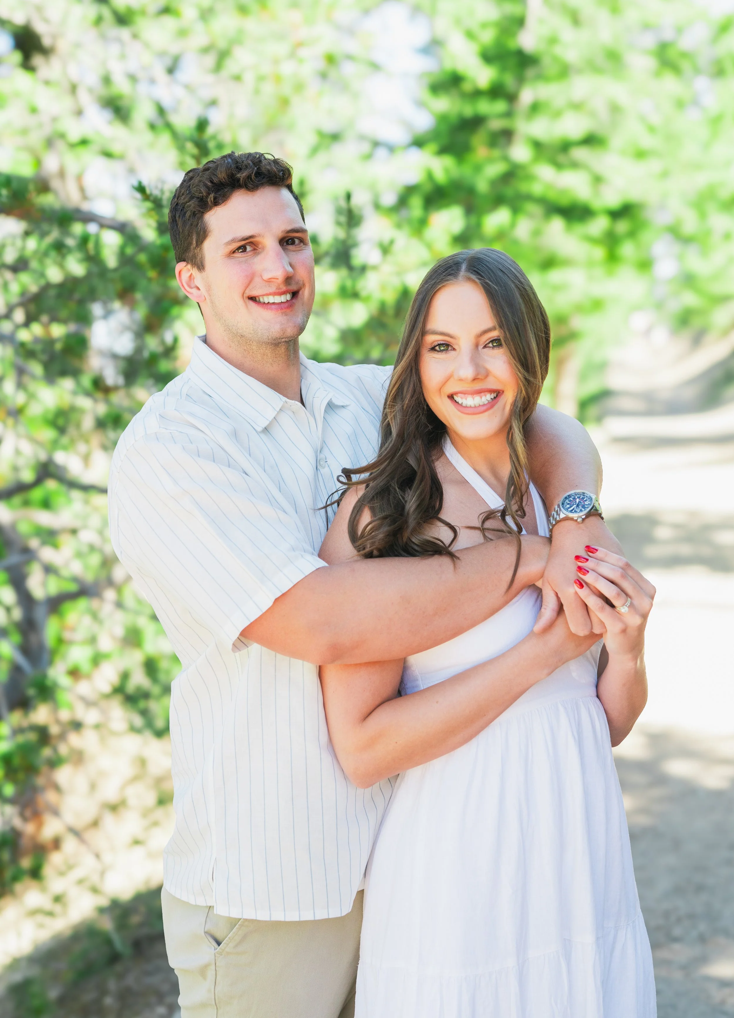 A happy couple standing outdoors on a sunny day, smiling and embracing each other. The man is wearing a white striped shirt and khaki pants, and the woman is in a white dress with long dark hair.