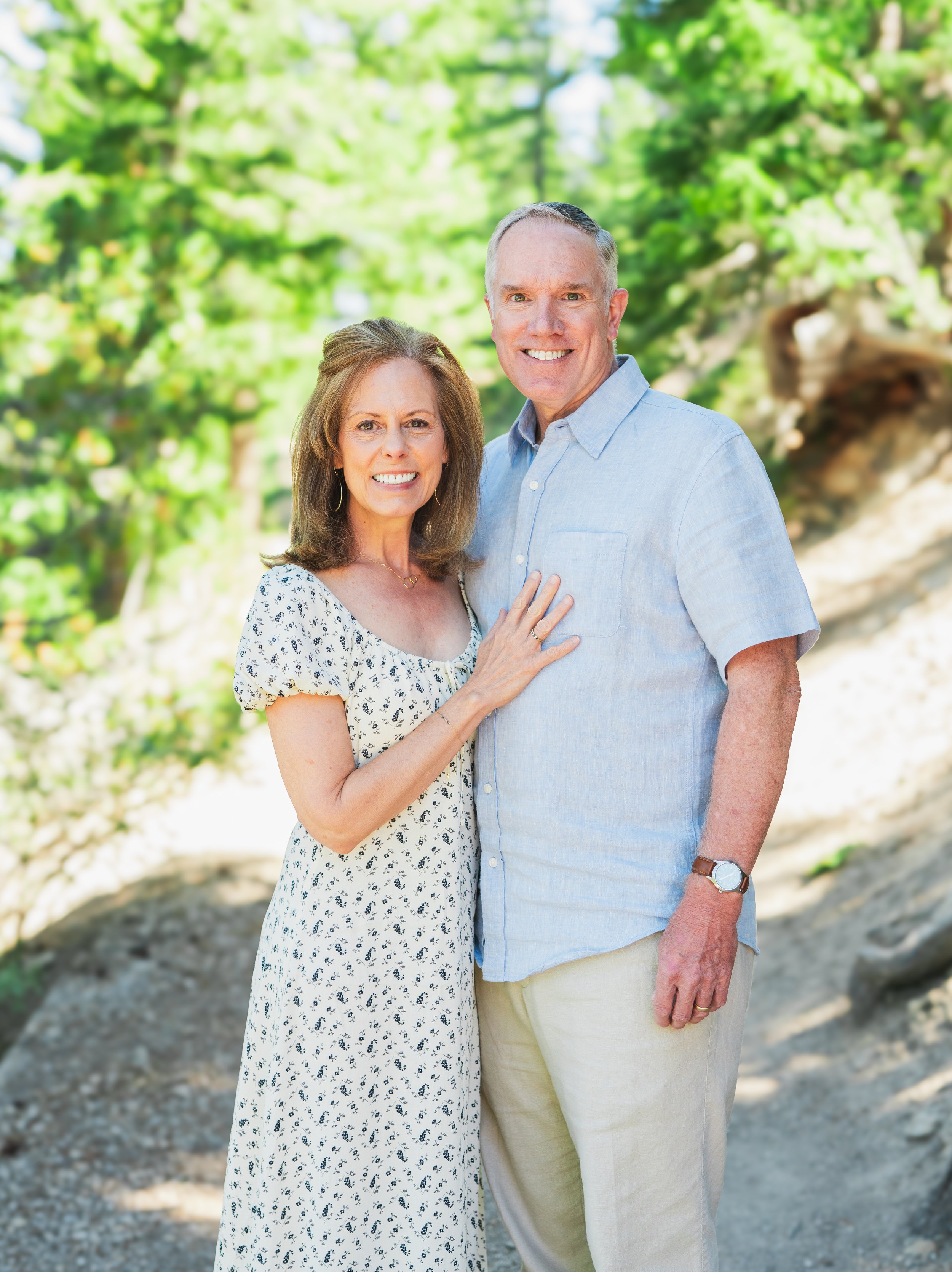 A couple smiling outdoors on a sunny day with green trees in the background. The woman is wearing a white floral dress and the man is wearing a light blue shirt and beige pants.
