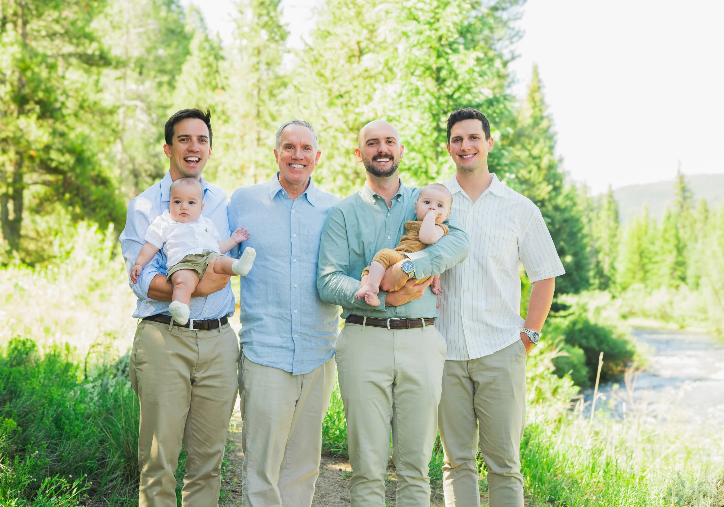 Group of five men, two holding babies, standing outdoors in a lush green park or forest, smiling at the camera.