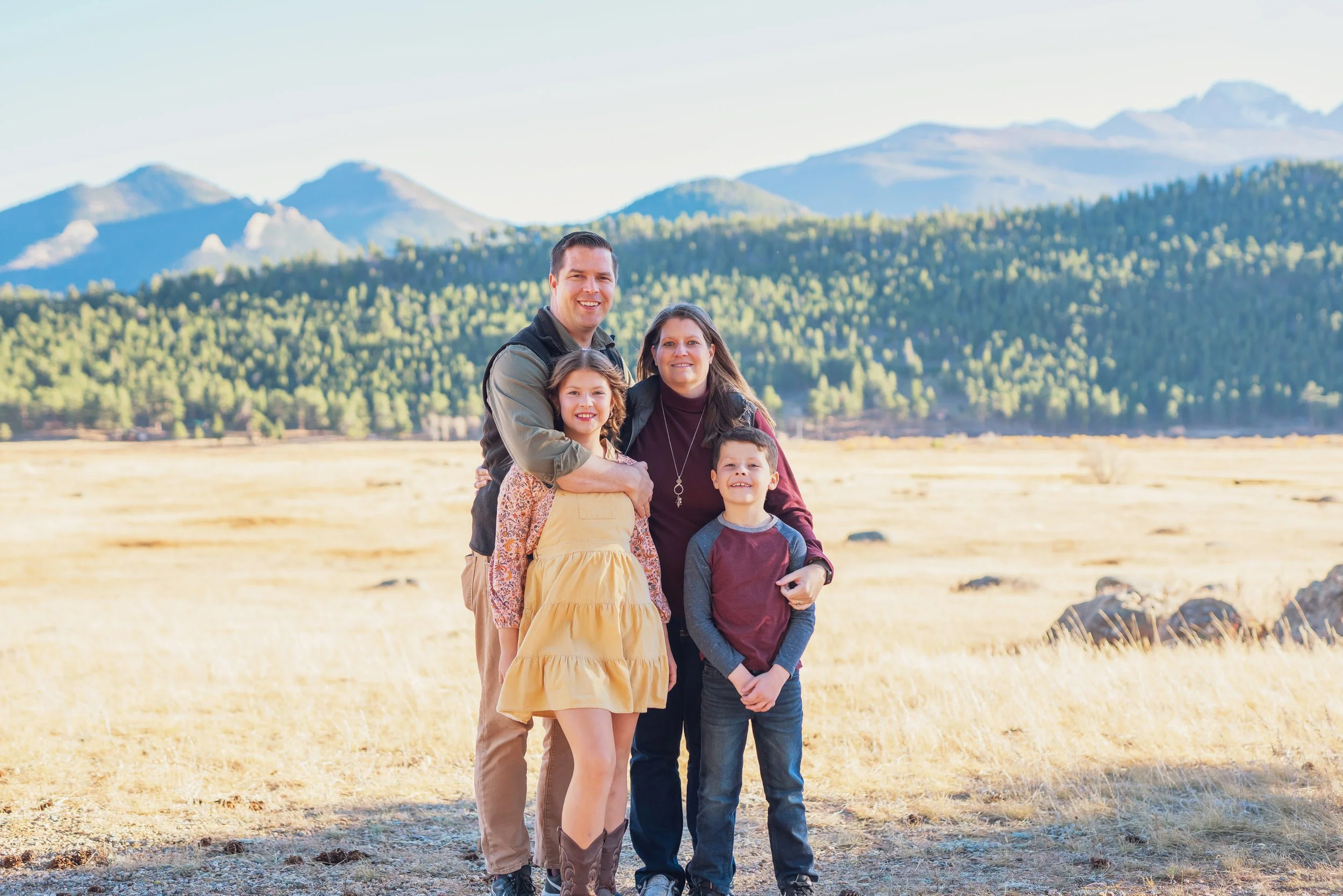 A family of four standing outdoors in a grassy field with mountains and green trees in the background, smiling at the camera.
