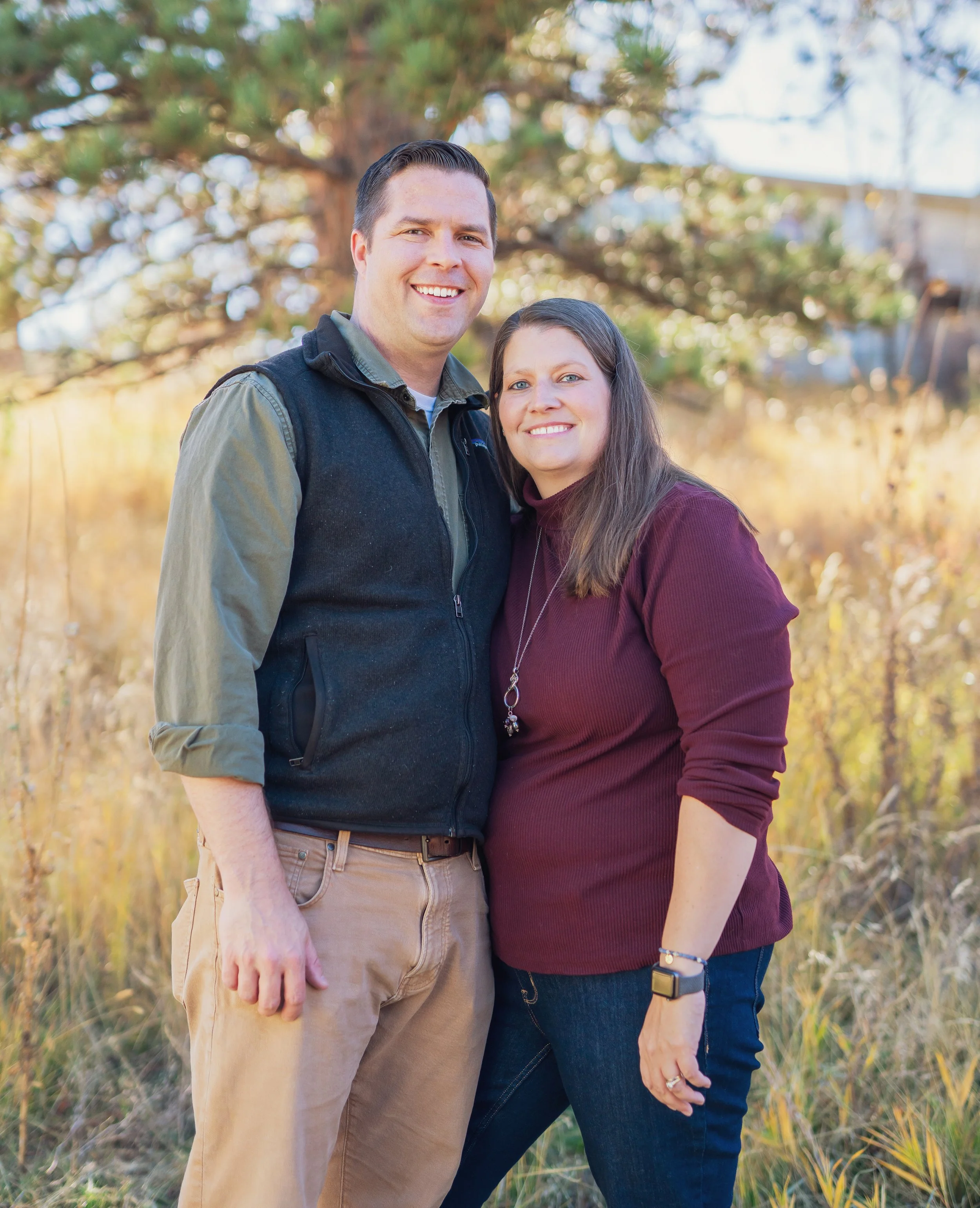 A smiling couple standing outdoors in a grassy area with trees in the background on a clear day.