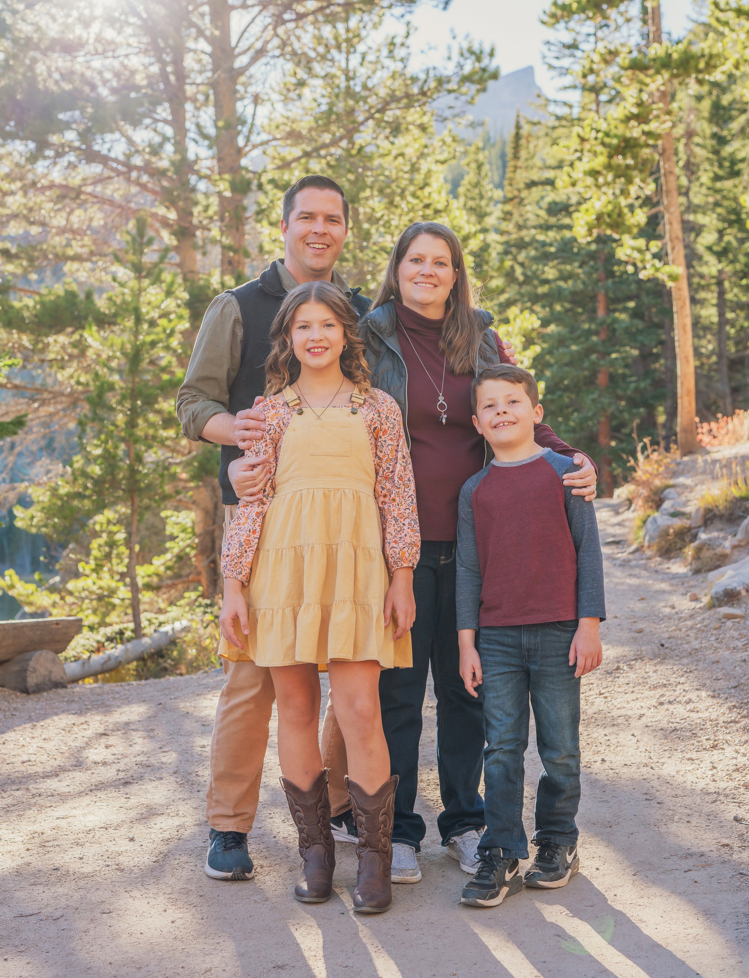 A family of five standing outdoors on a dirt path in a forested area with trees and sunlight in the background.