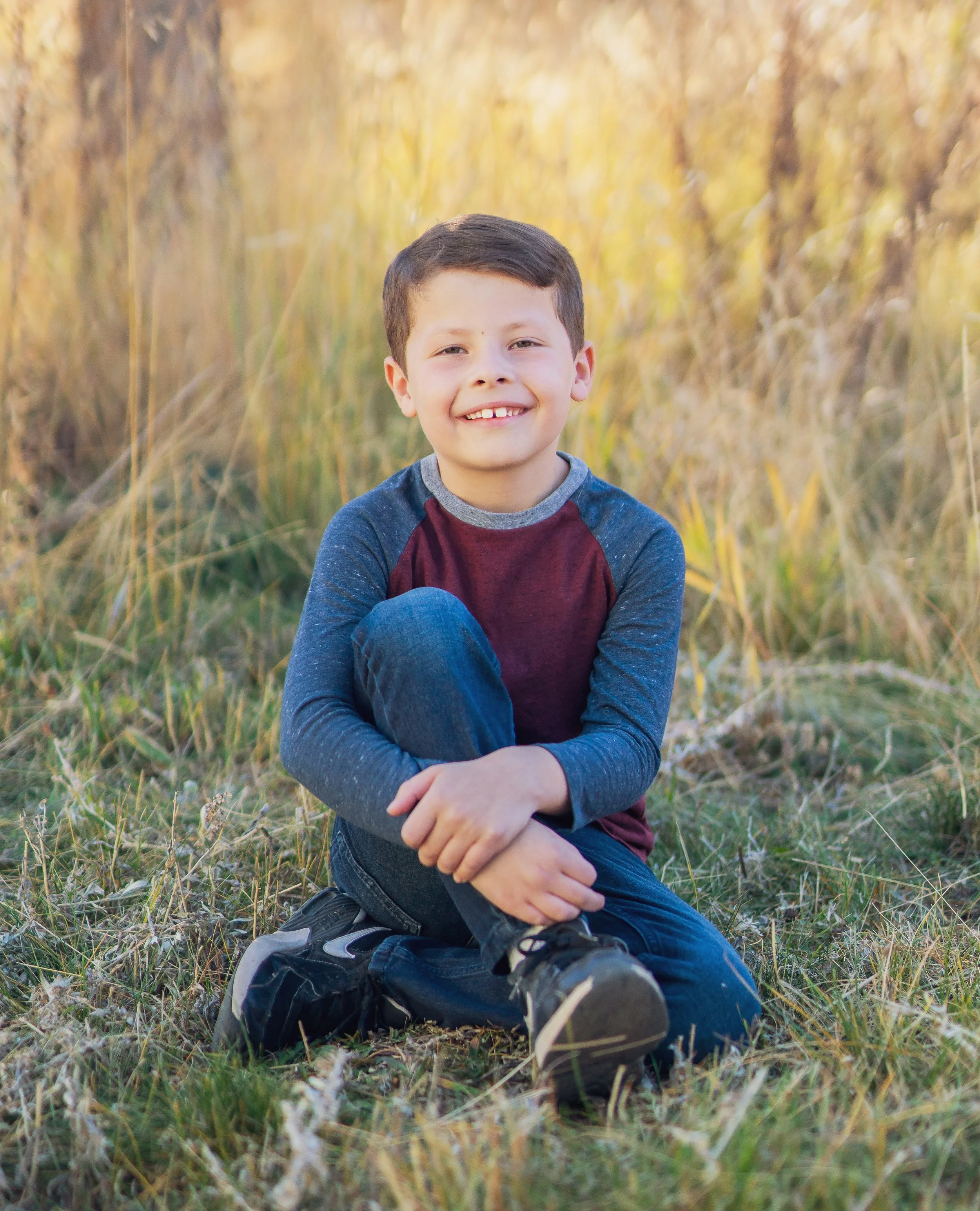 A young boy sitting cross-legged in a field of tall grass, smiling at the camera during golden hour.