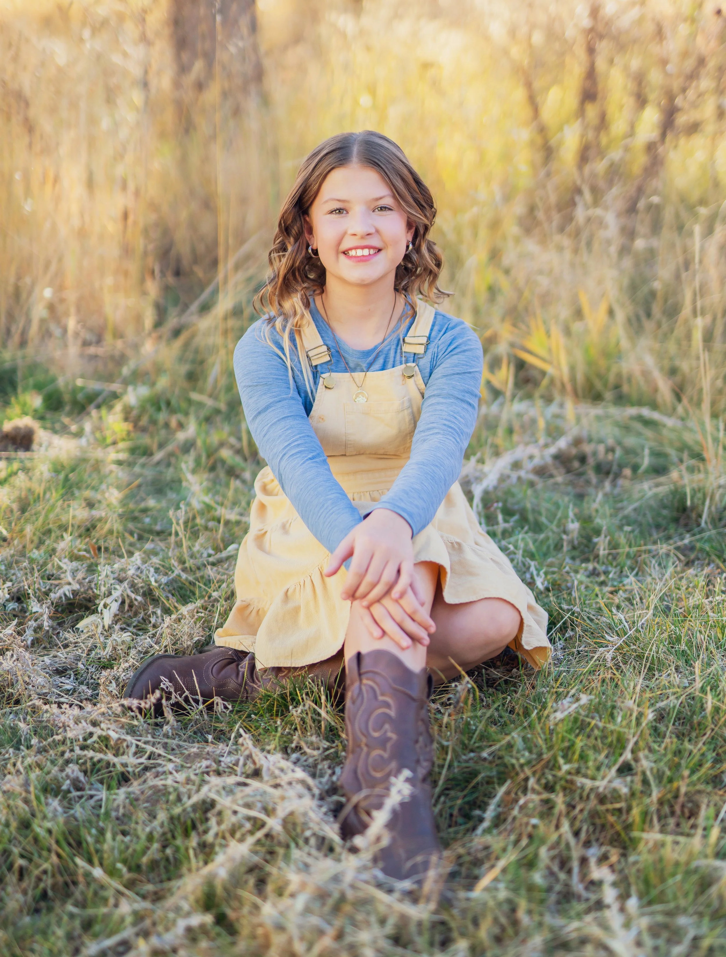A young girl with wavy brown hair, smiling, sitting on the grass in an autumn field. She is wearing a blue long-sleeve shirt, a yellow dress, and cowboy boots. The background is filled with tall yellow and green grass.