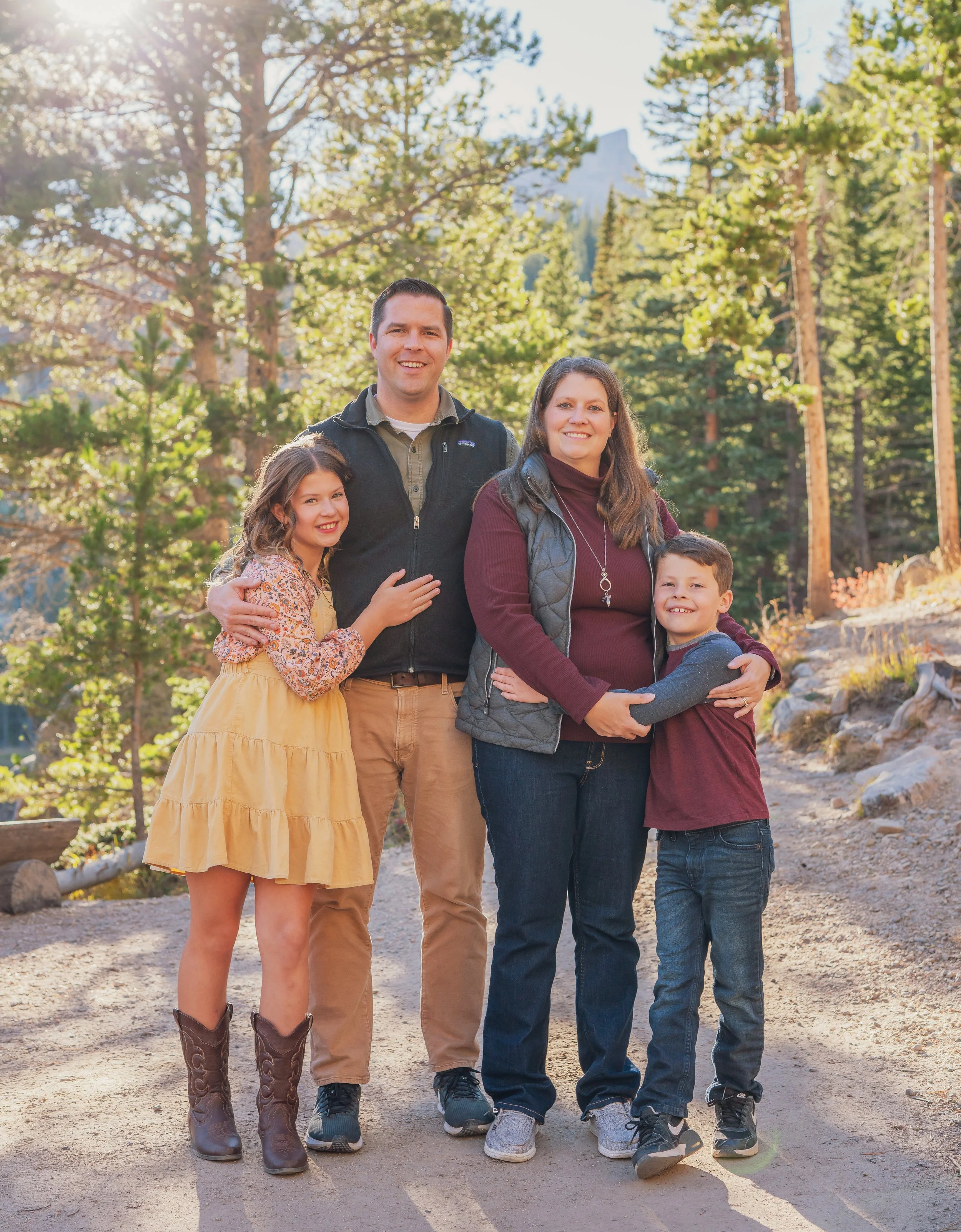 A family of four, including a man, a woman, a teenage girl, and a young boy, standing outdoors in a wooded area with trees and mountains in the background on a sunny day.