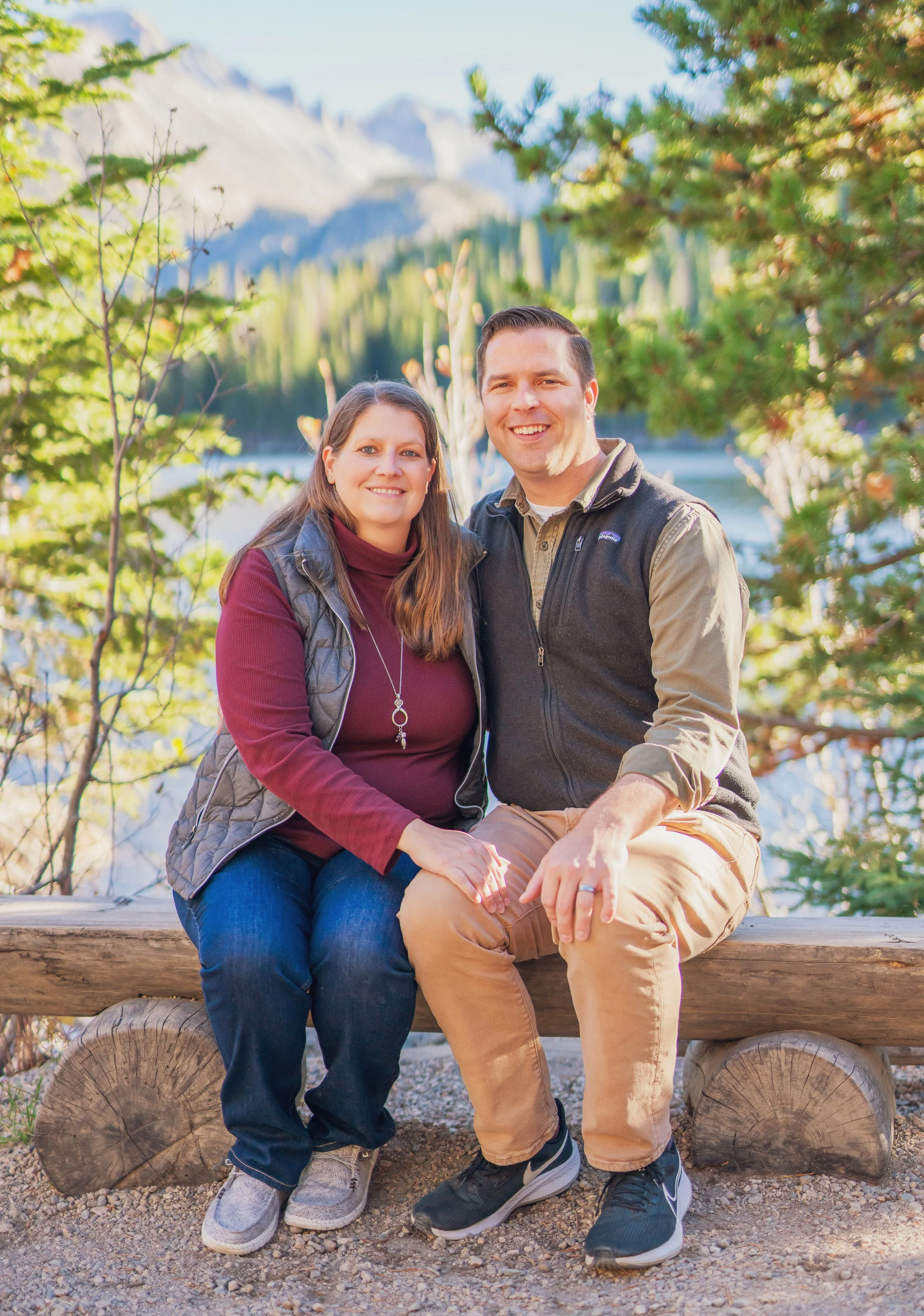 A smiling couple sitting on a log bench outdoors, surrounded by trees, with mountains and a lake in the background during daytime.