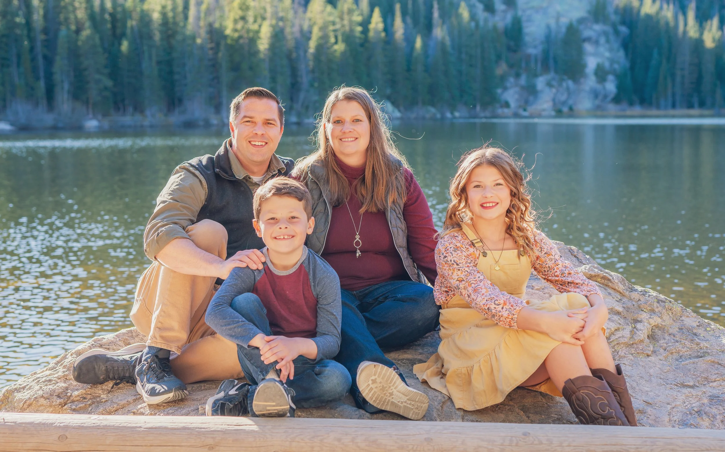 Family of five sitting on a rock near a lake with trees in the background, smiling at the camera.