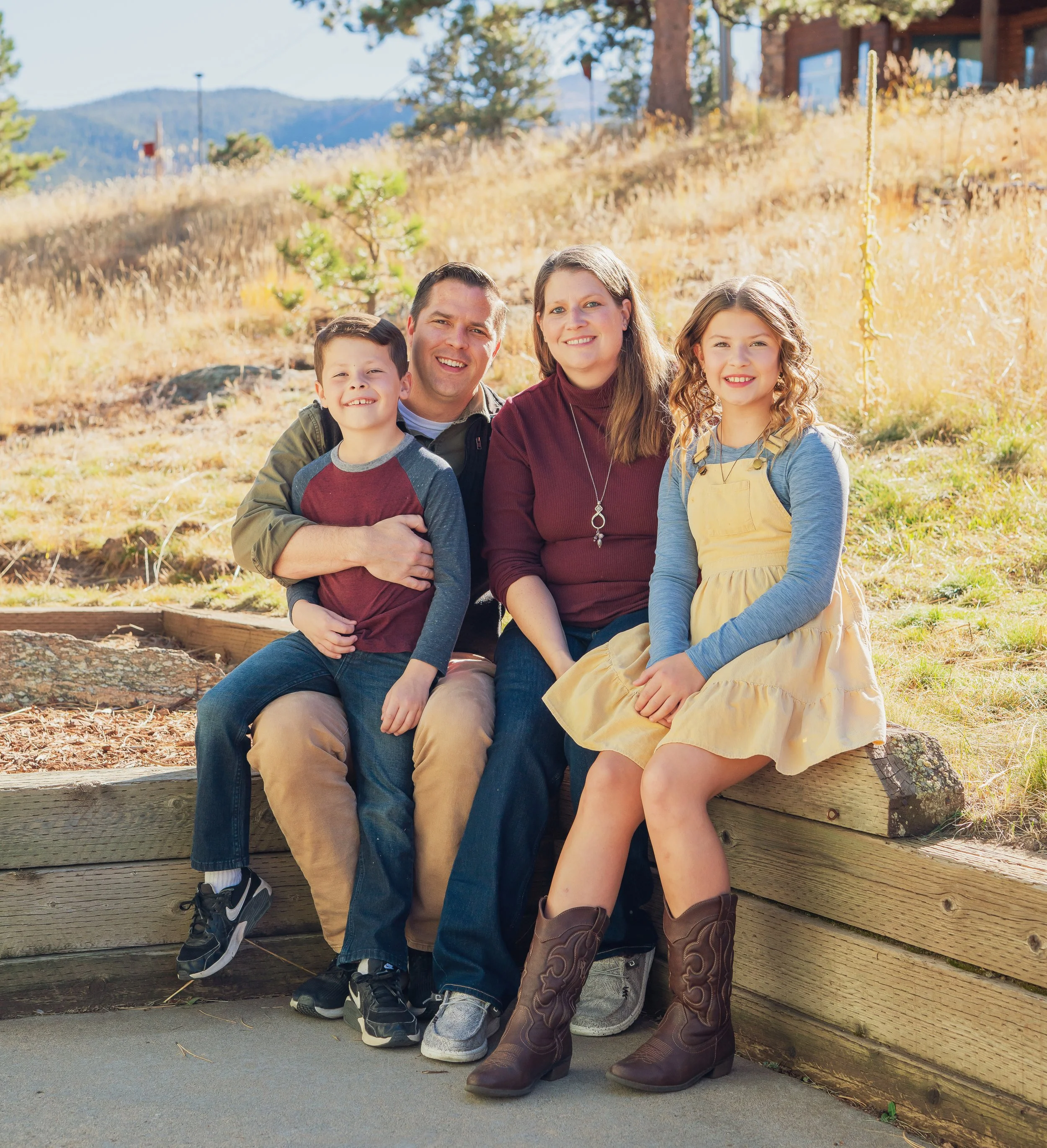 A family of four sitting outdoors on a wooden retaining wall, smiling at the camera during fall, with trees and hills in the background.