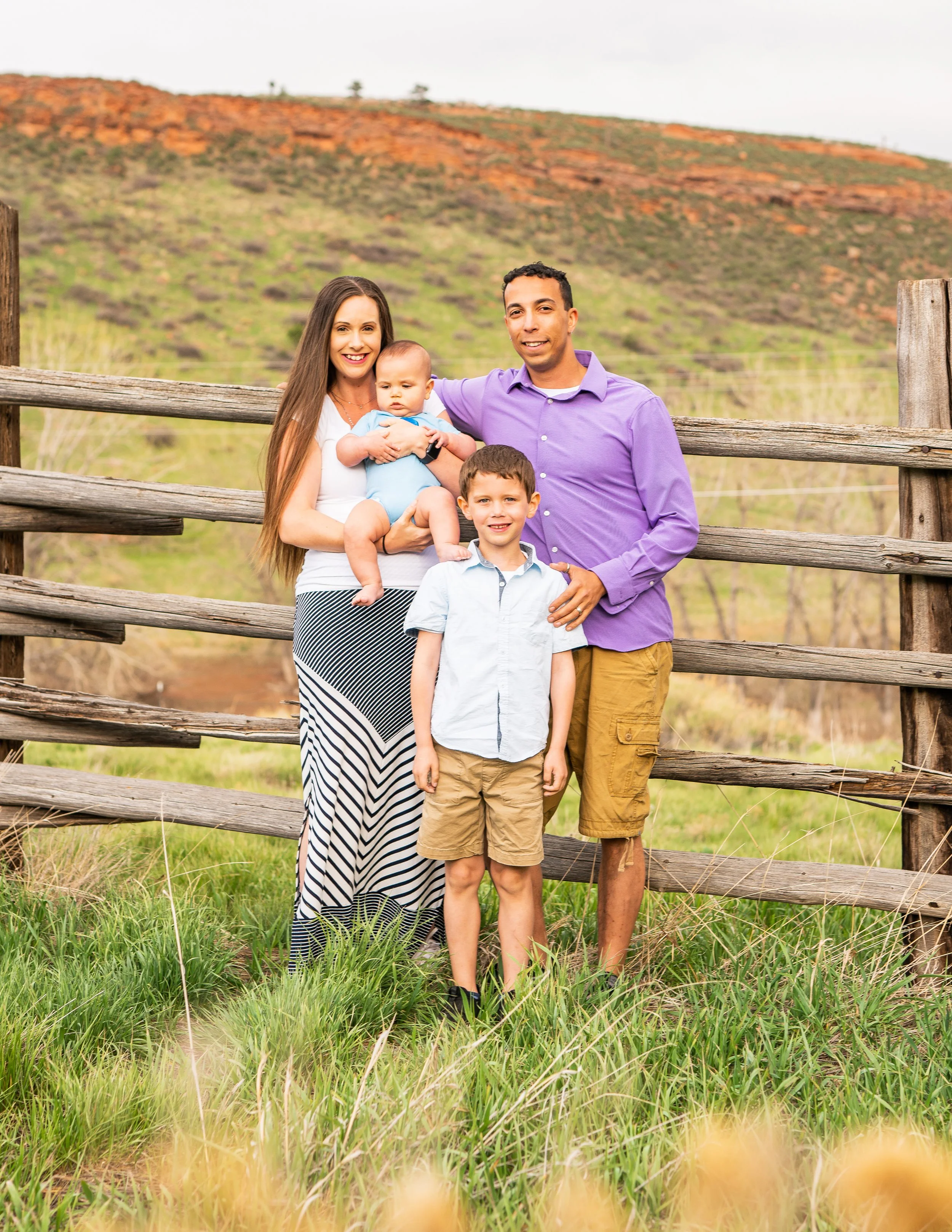 Family of four posing outdoors near a wooden fence with a grassy field and a hill in the background