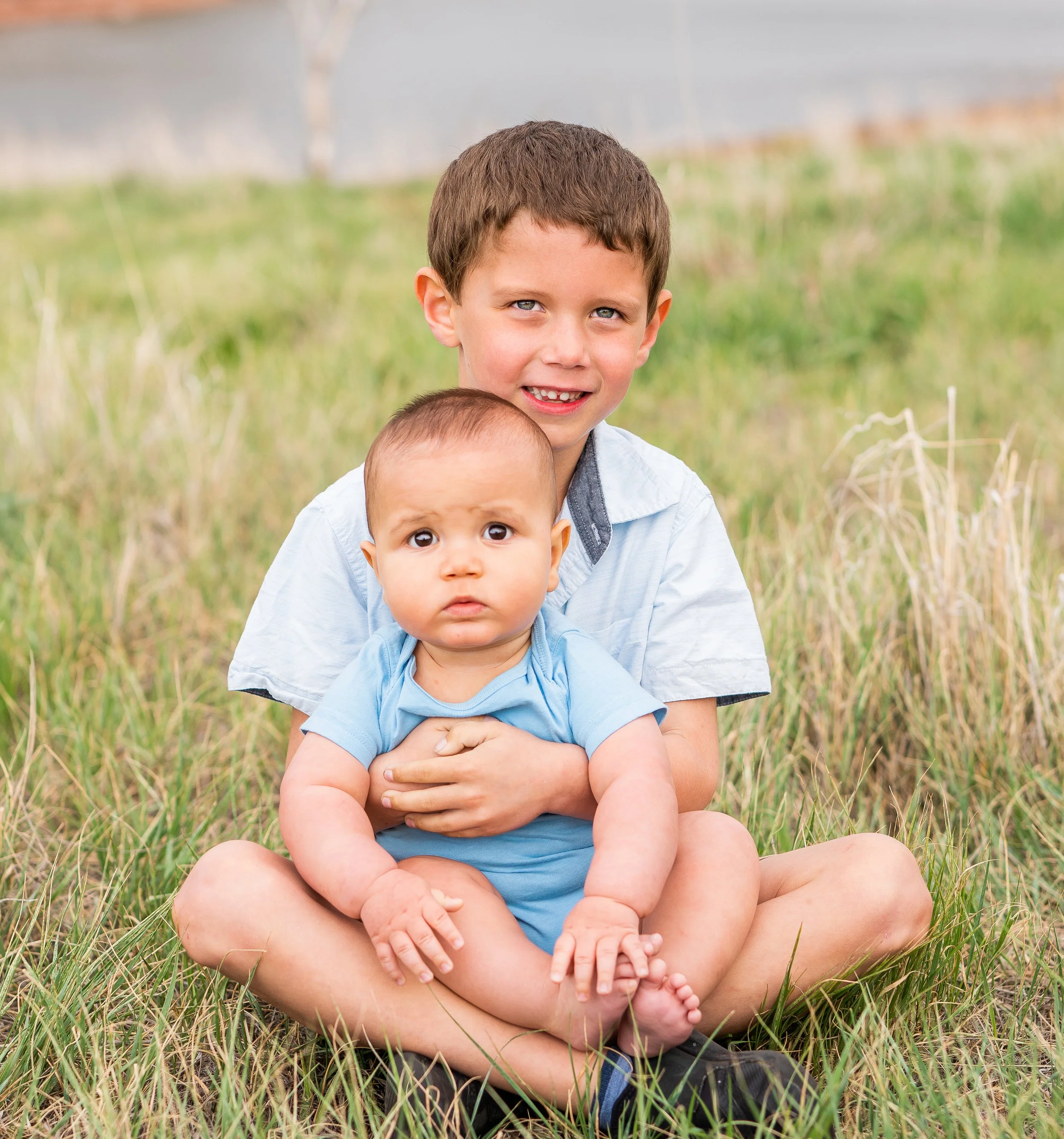 Two young children, an older boy and a younger baby, sitting on grass outdoors with the older boy holding the baby in his lap. The boy is smiling slightly, and the baby has a serious expression. Background shows a field with some greenery.