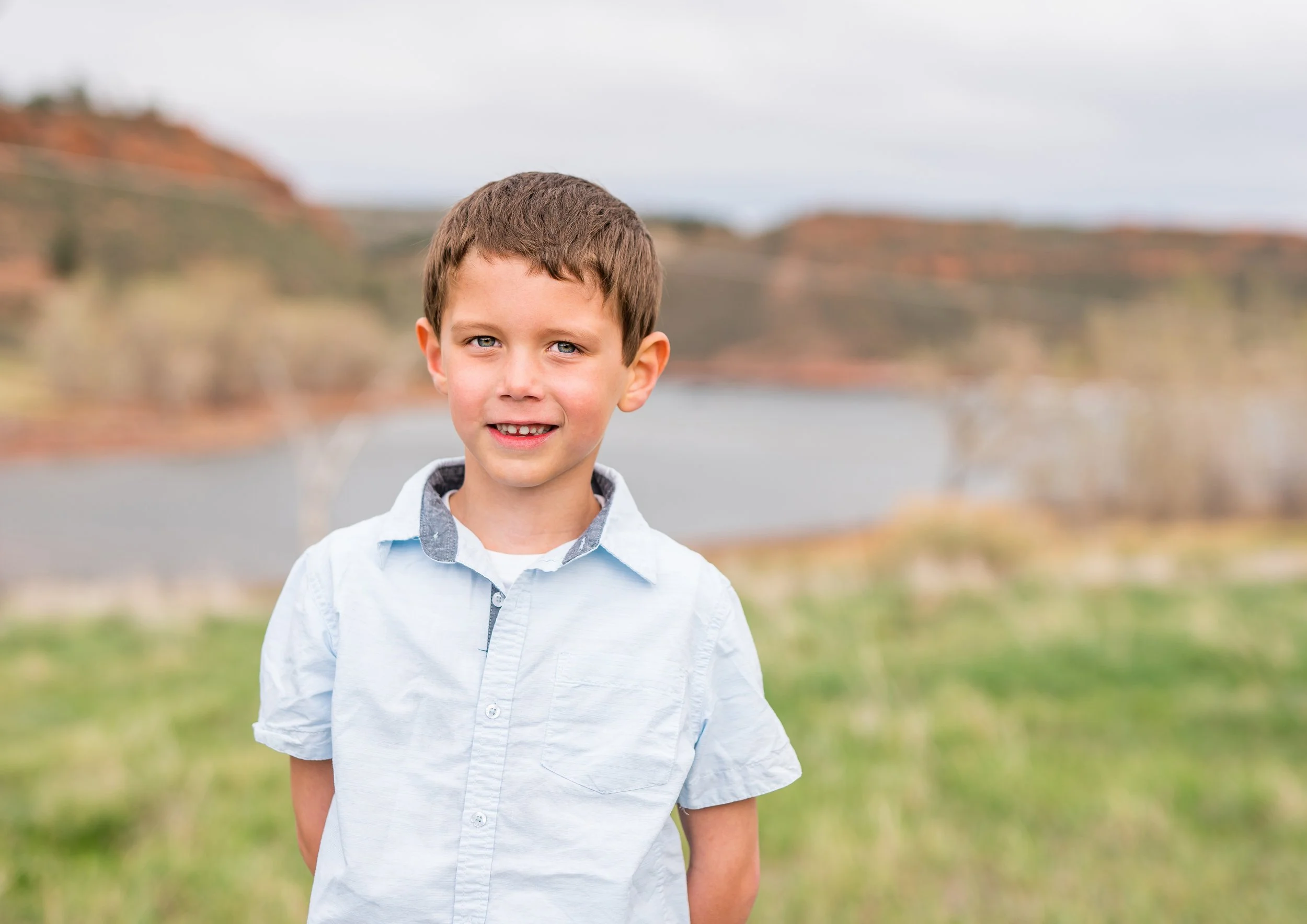 A young boy with short brown hair and blue eyes, wearing a light blue shirt, stands outdoors with a blurred lake and hills in the background.
