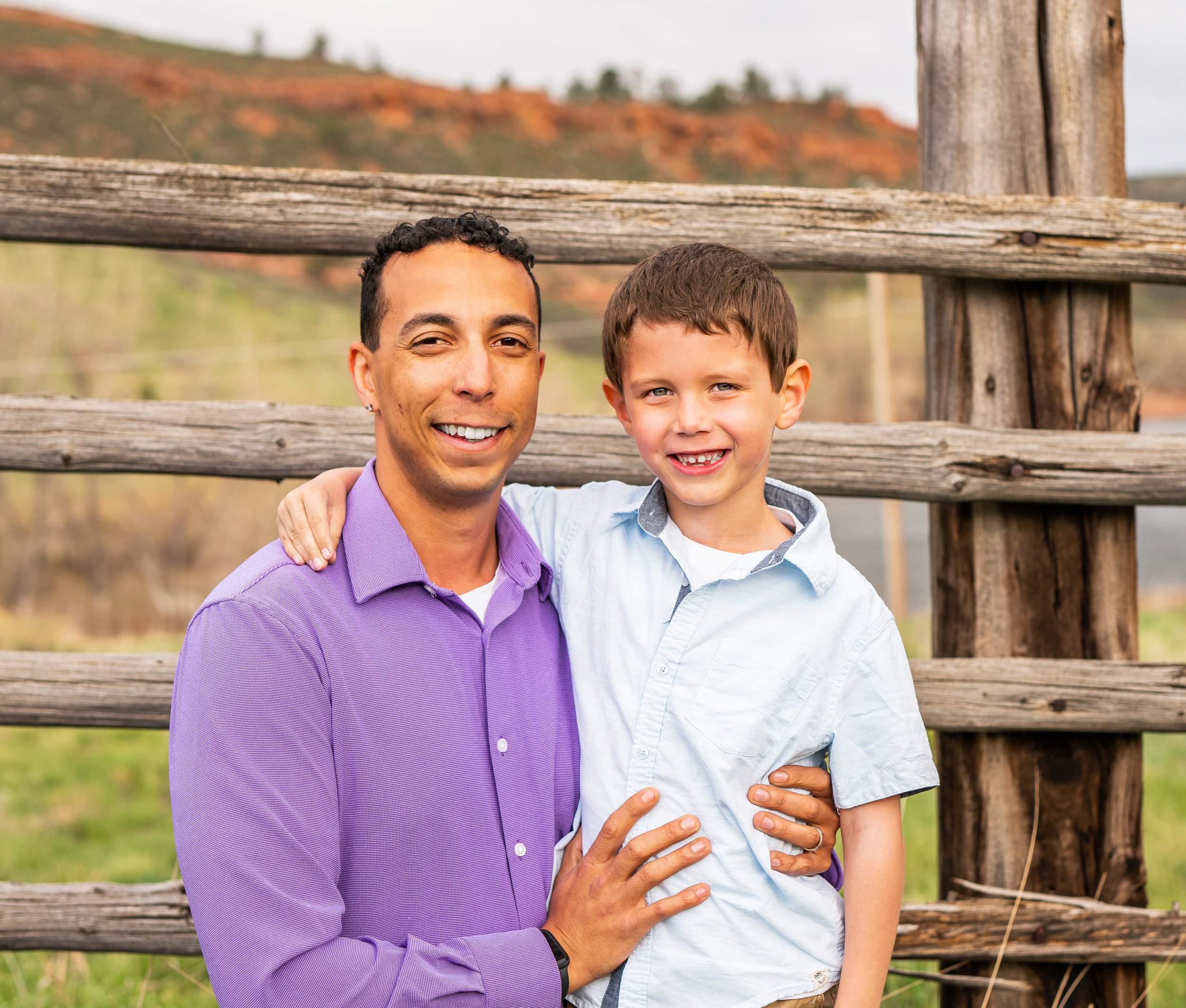 A man and young boy smiling and embracing outdoors in front of a wooden fence with trees and a hill in the background.