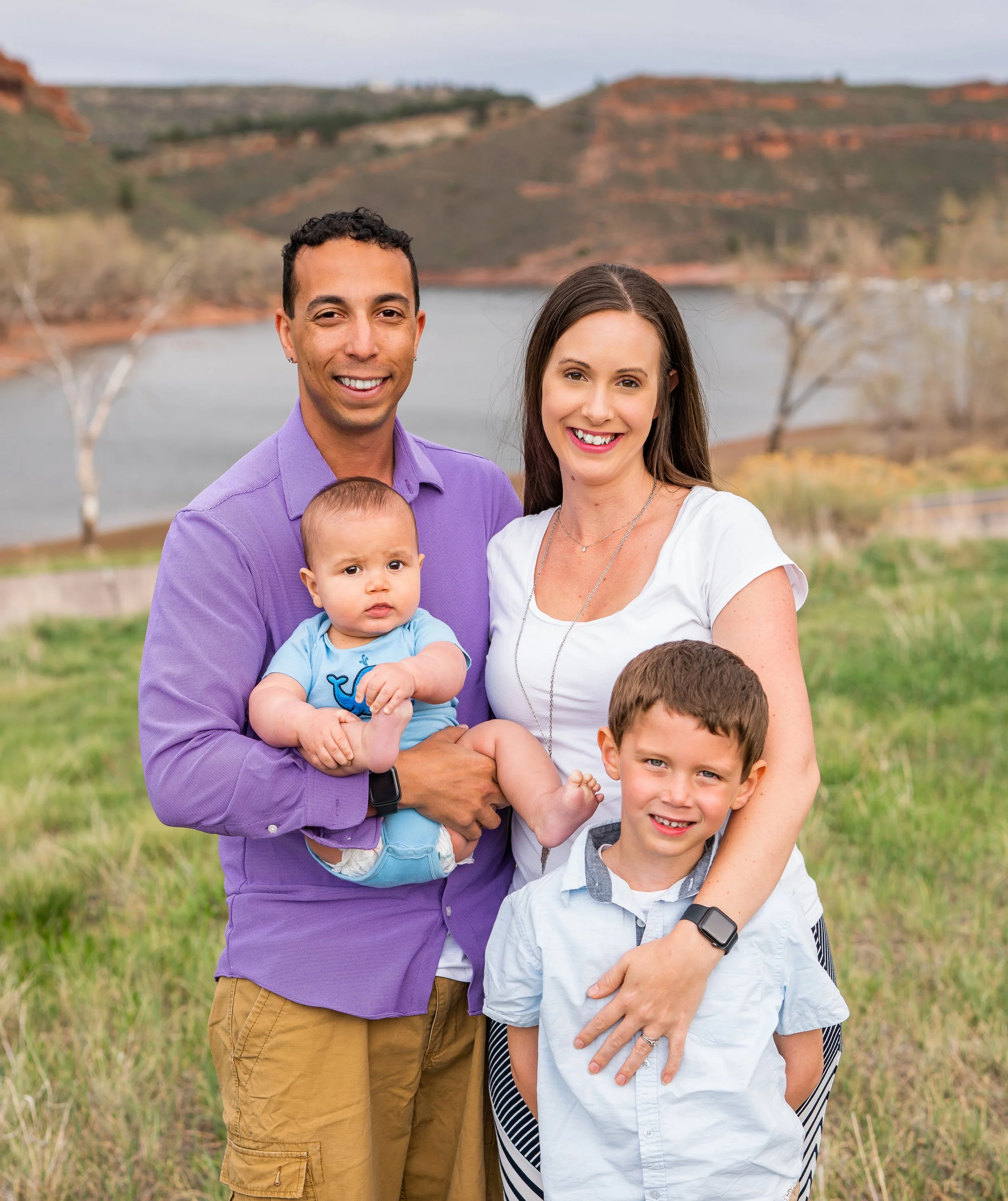 A family of four standing outdoors near a lake with hills in the background, smiling at the camera. The father, wearing a purple shirt, holds a baby in a blue outfit, while the mother, in a white top, stands beside a young boy in a white shirt.