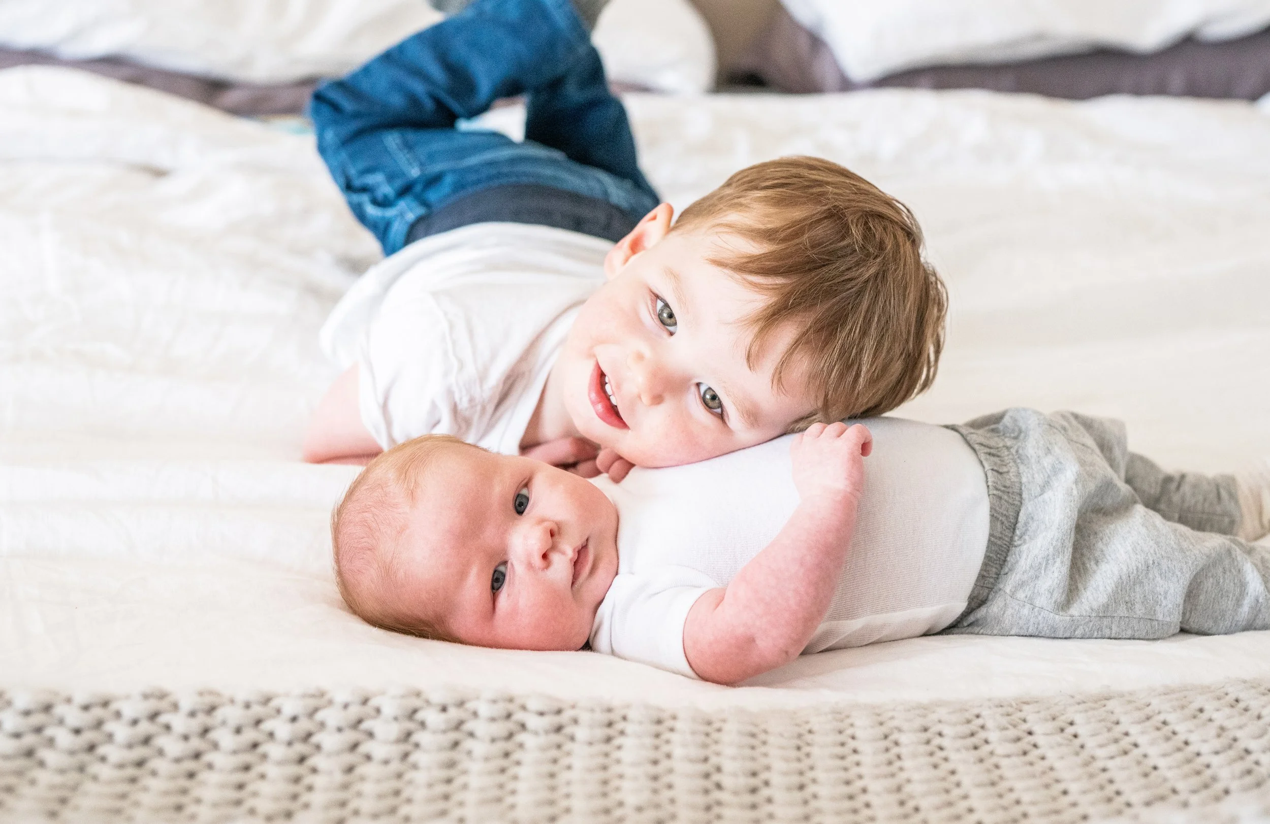 A young boy lying on a bed with his head resting on a baby, both looking at the camera.