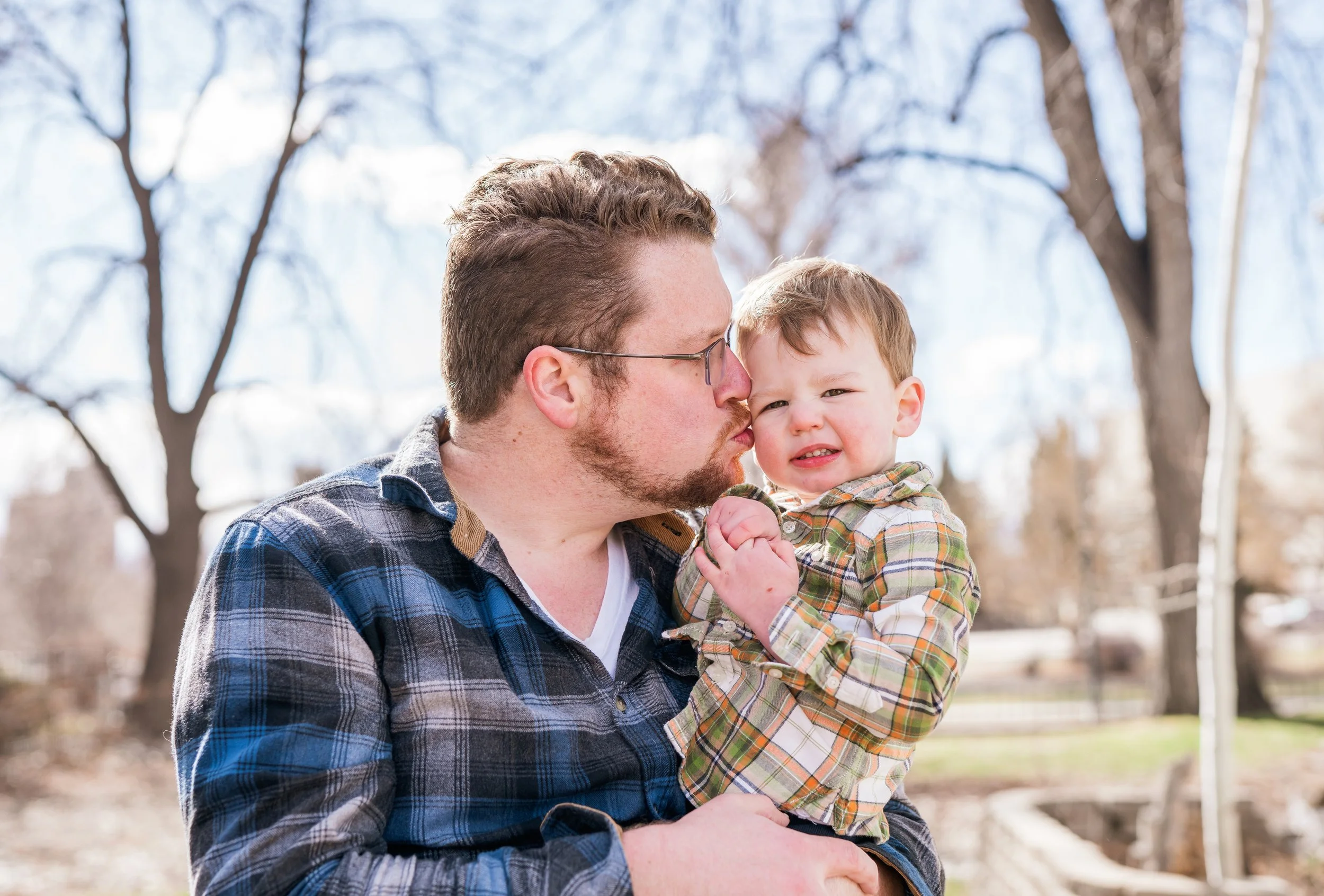 A man with glasses kisses a young boy on the face outdoors, both wearing plaid shirts. The boy looks uncomfortable. Trees and a park setting are in the background.