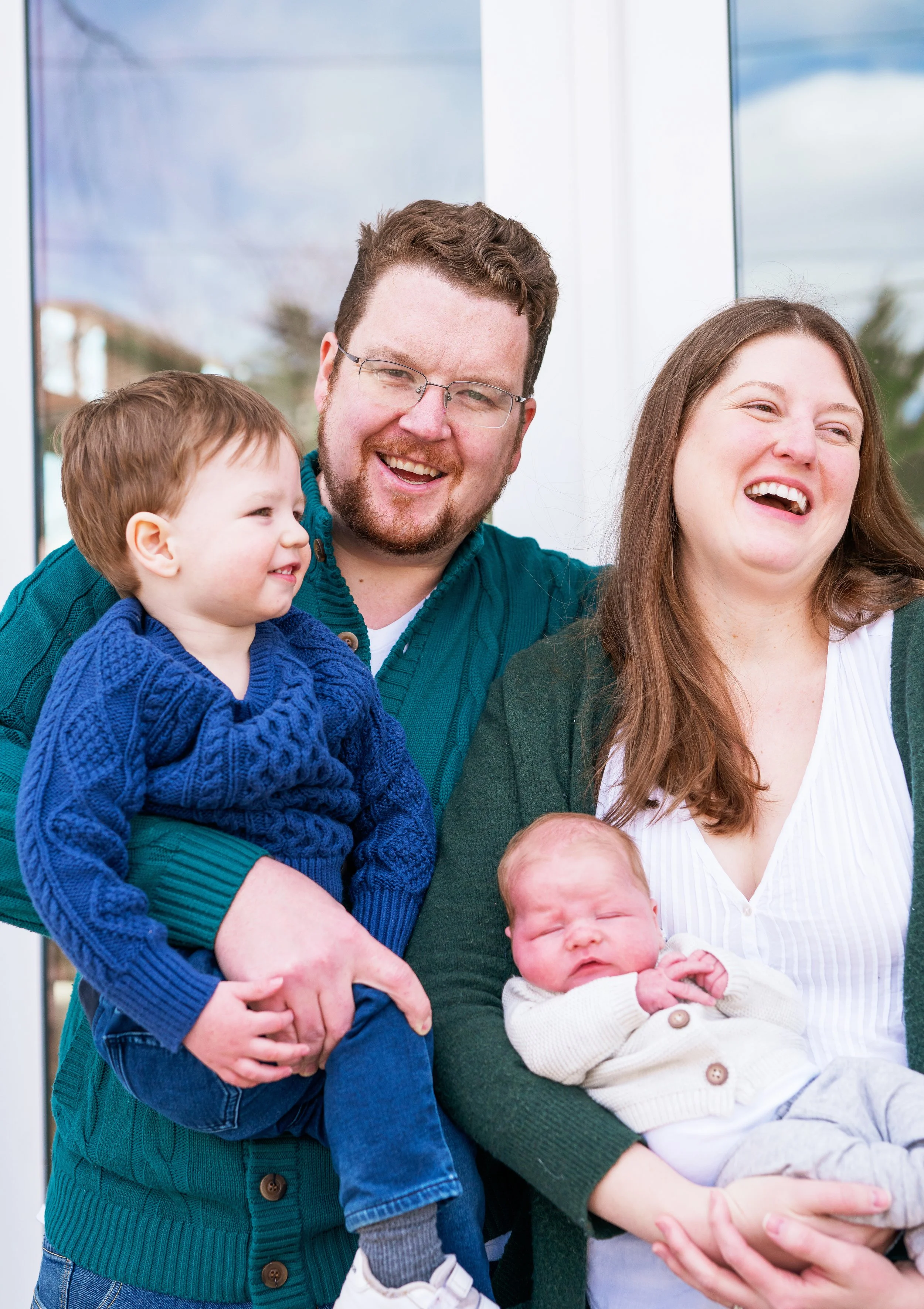A smiling family of four, including a man, woman, and two young children, laughing together in a bright indoor setting.