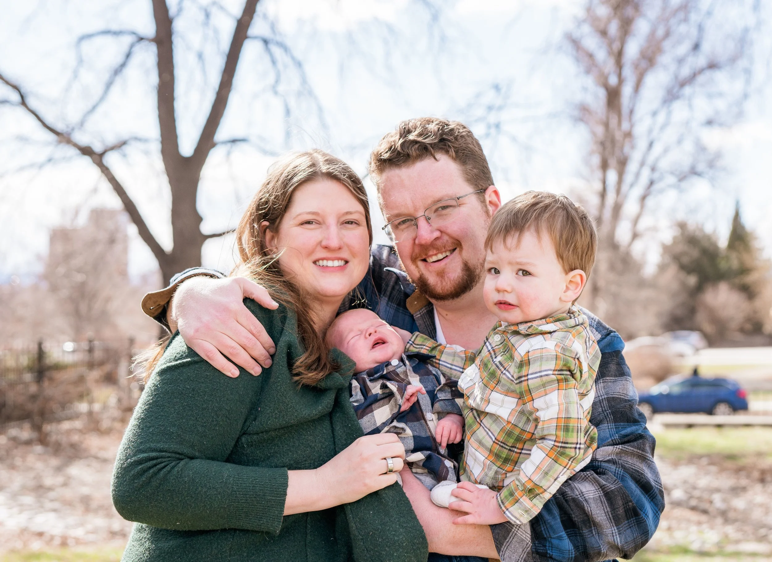A family of four outdoors in a park during daytime. The mother, father, a young boy, and a newborn baby are smiling and holding each other.