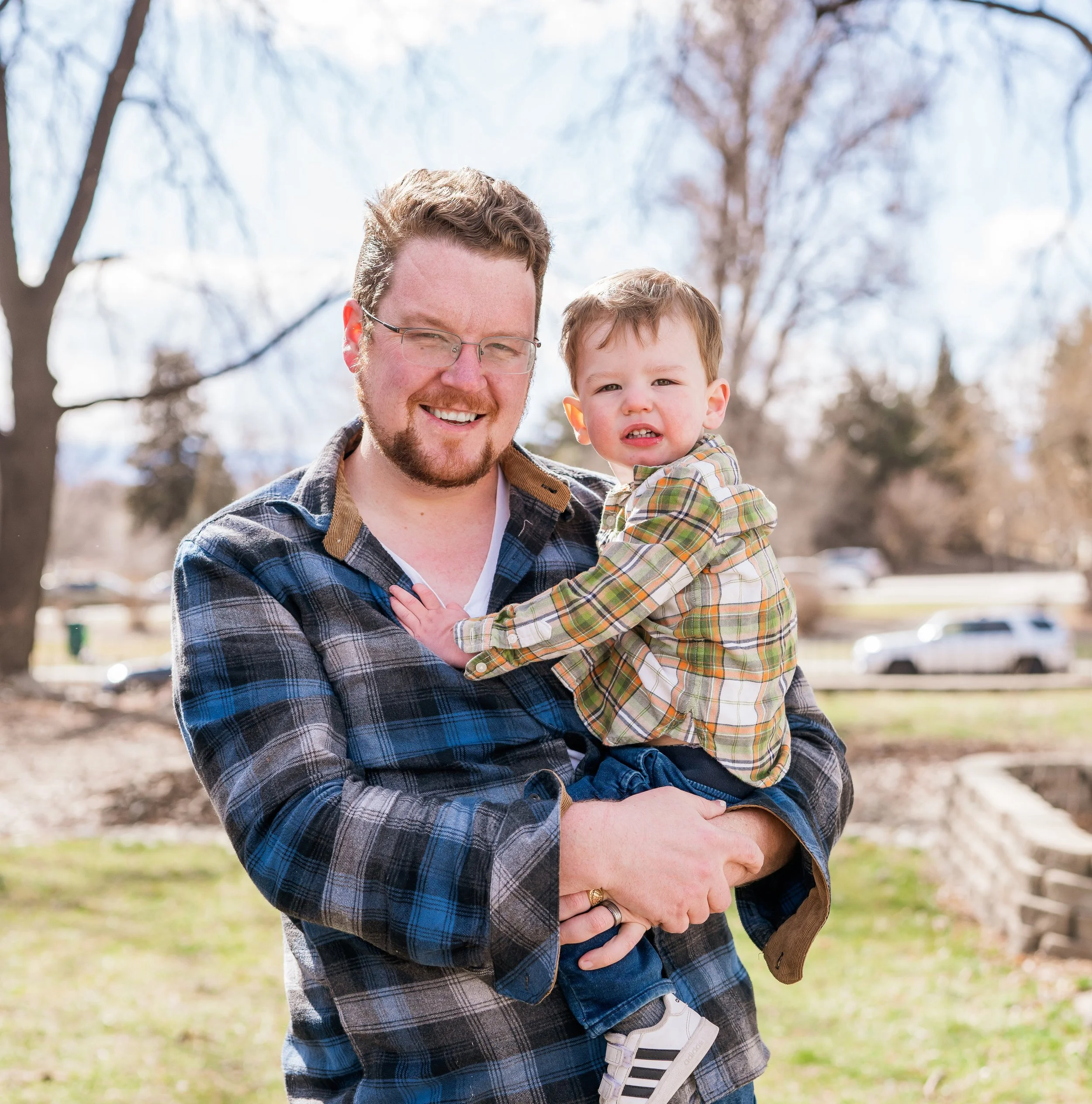 A man with glasses and a beard smiling while holding a young boy in an outdoor park on a sunny day.