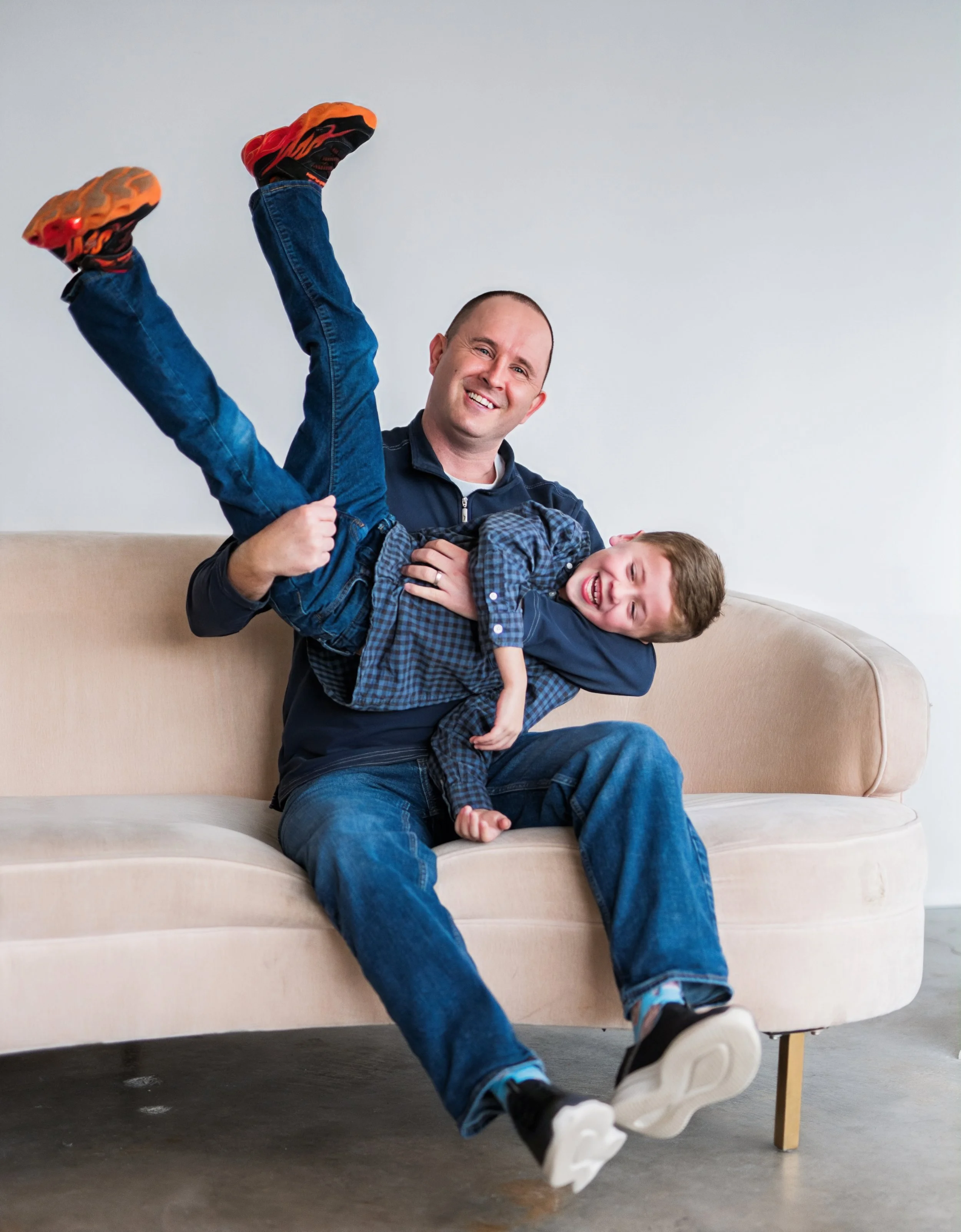 A man sitting on a beige sofa holding a laughing boy in his lap and lifting him up, both wearing casual clothes and smiling.