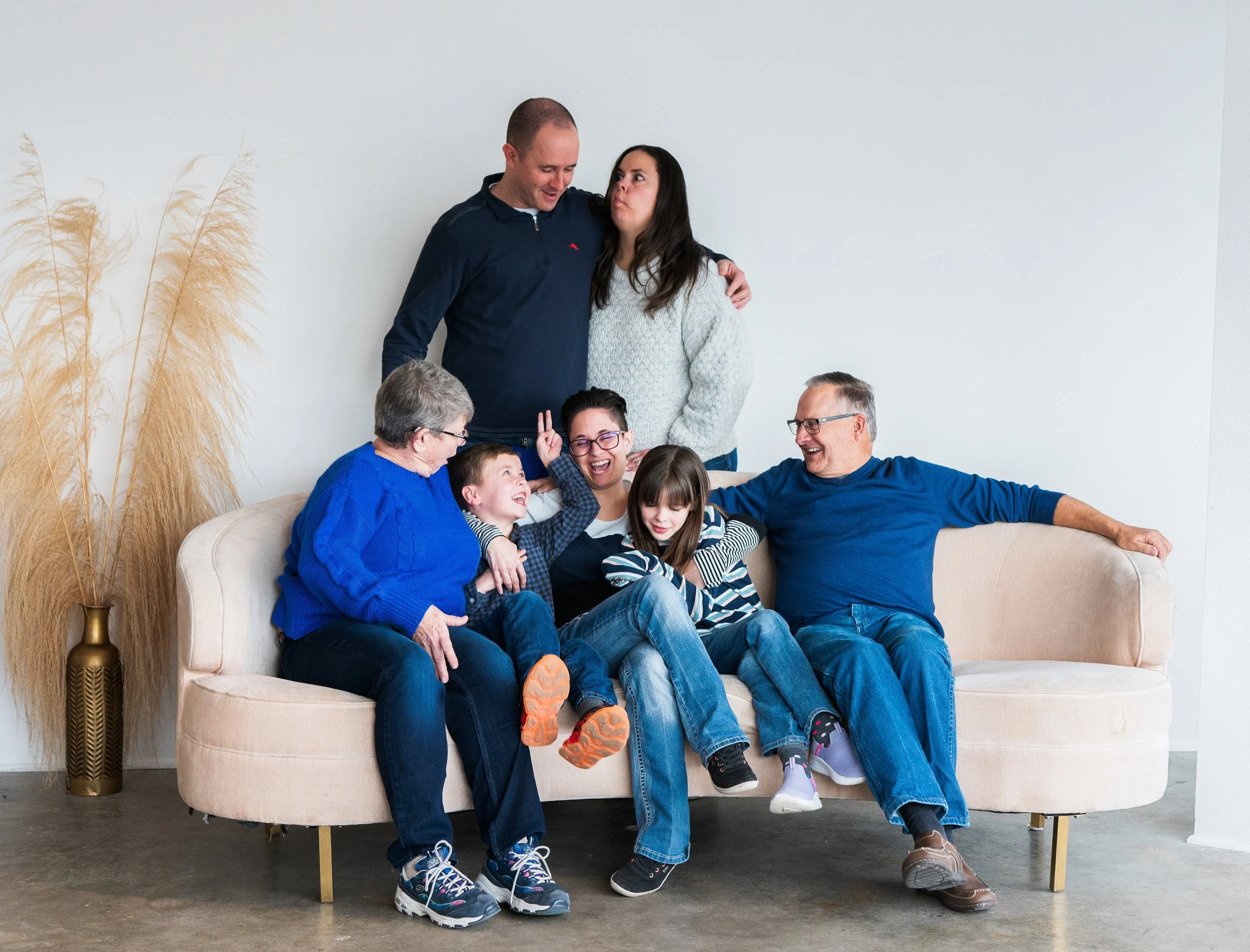 Family of seven sitting and standing on and around a light-colored sofa in a living room, laughing and enjoying each other's company.