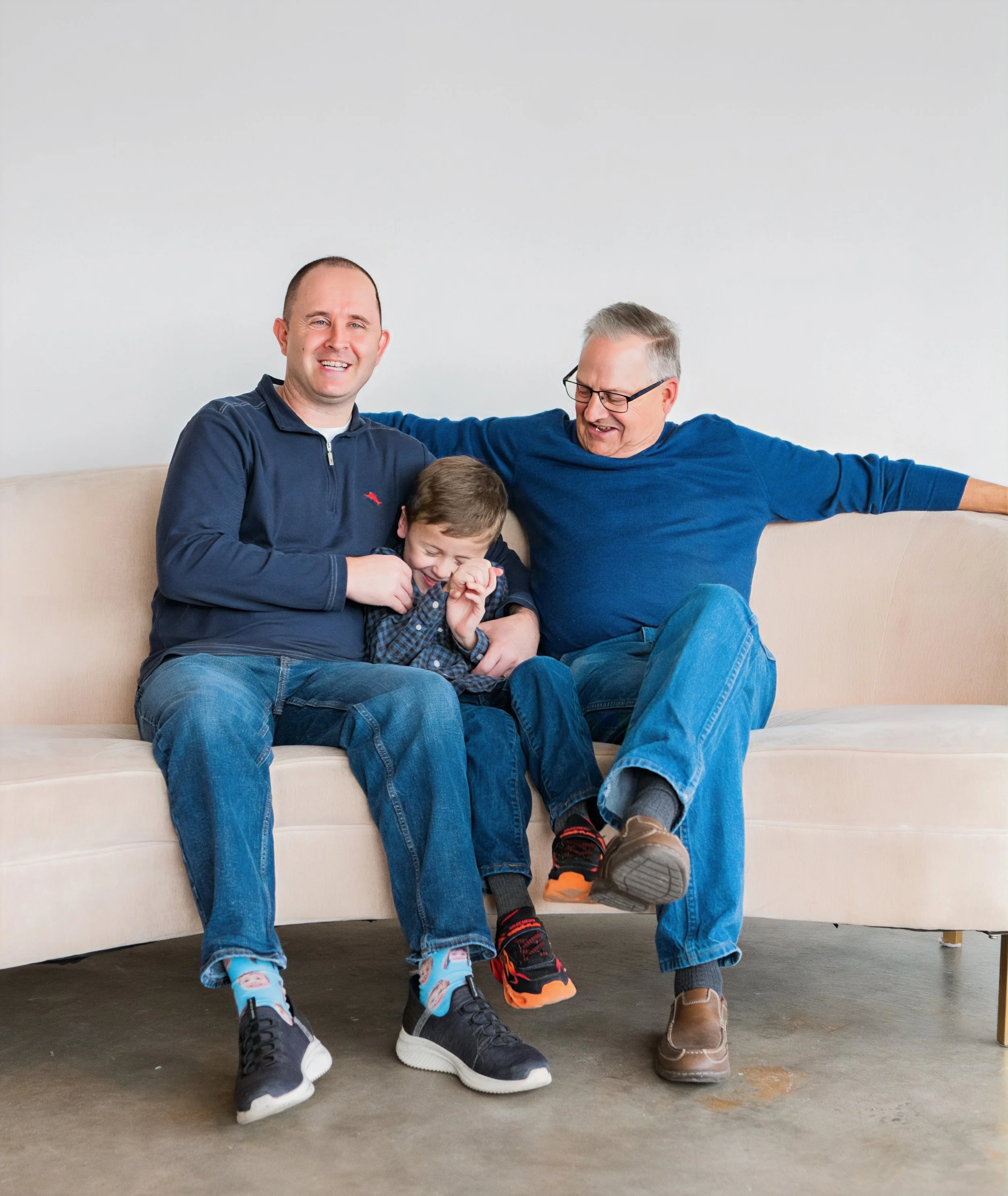 Three males sitting on a beige couch, laughing and enjoying each other's company, one of them is a young boy.