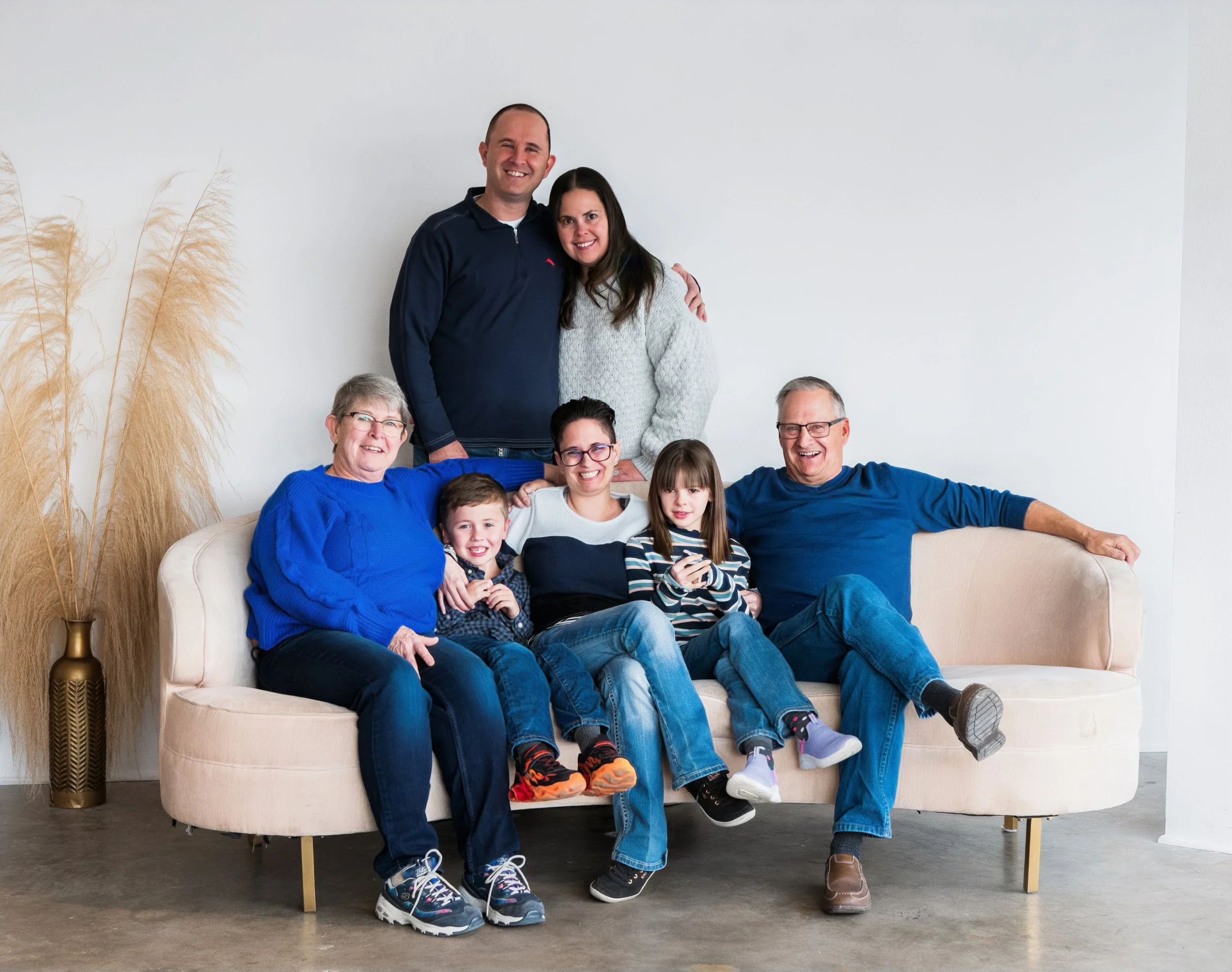 Family photo of seven people sitting and standing on a beige sofa in a bright room, with a tall vase containing dried pampas grass on the left side of the image.