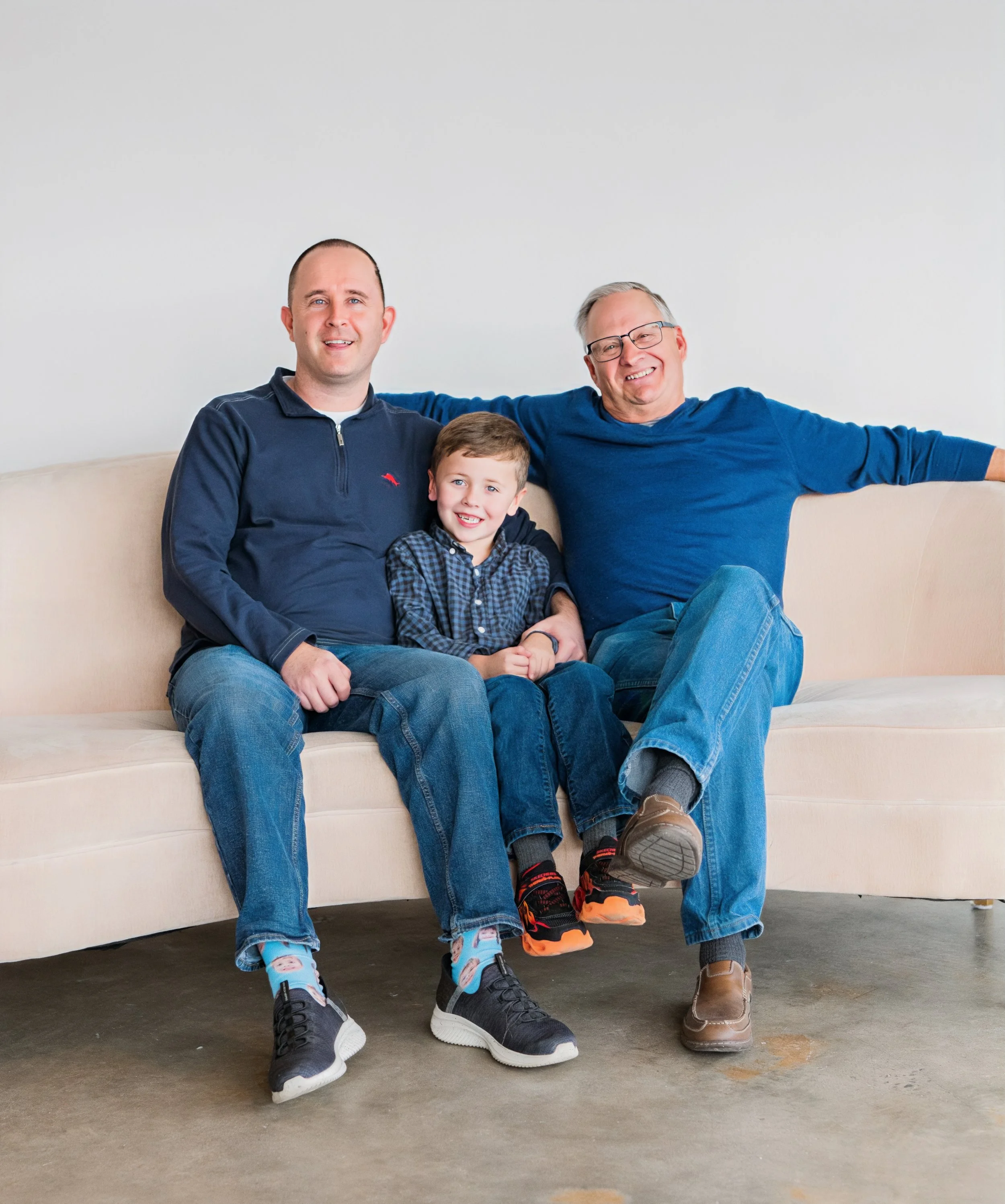 Three generations of a family sitting on a beige couch, smiling and relaxed. The man on the left has a shaved head and is wearing a navy blue zip-up sweater. The boy in the middle has short brown hair and is wearing a black and blue checkered shirt. 