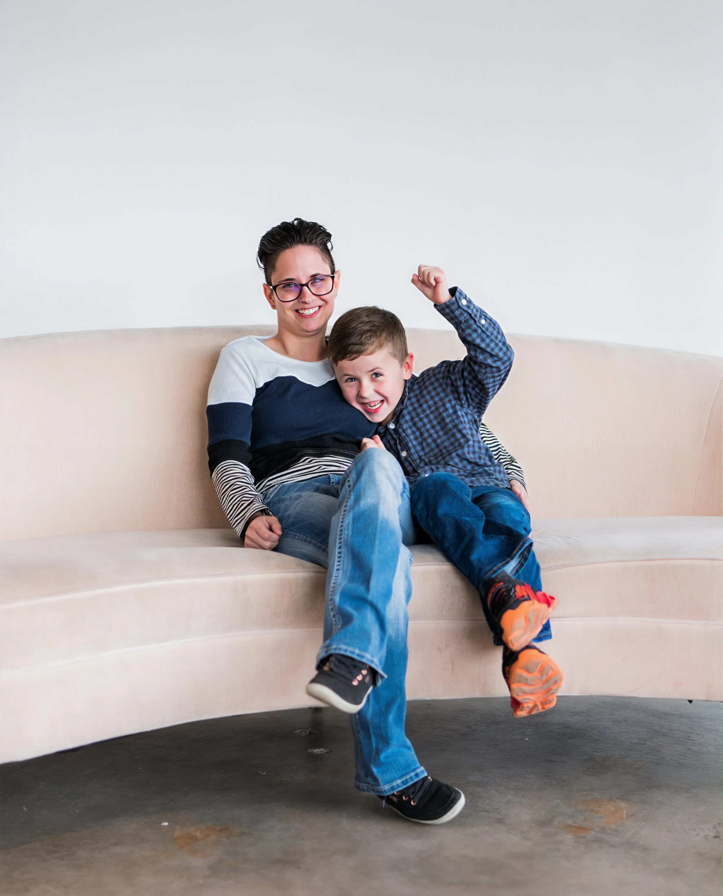 A woman and a young boy sitting on a beige couch, smiling and playing together, with the boy raising his arm in excitement.