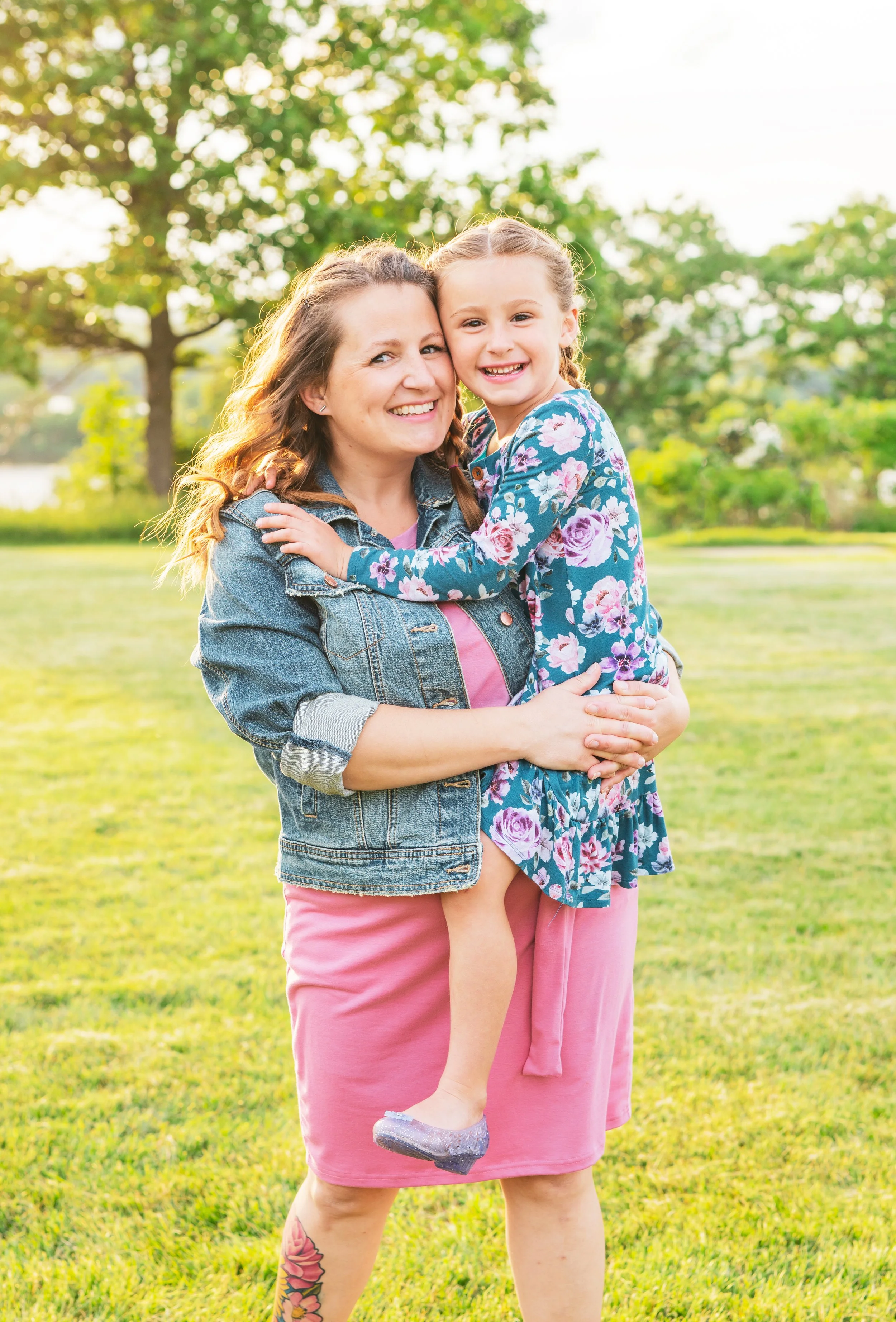 A woman and a young girl smiling and hugging outdoors in a park with green grass, trees, and sunlight.