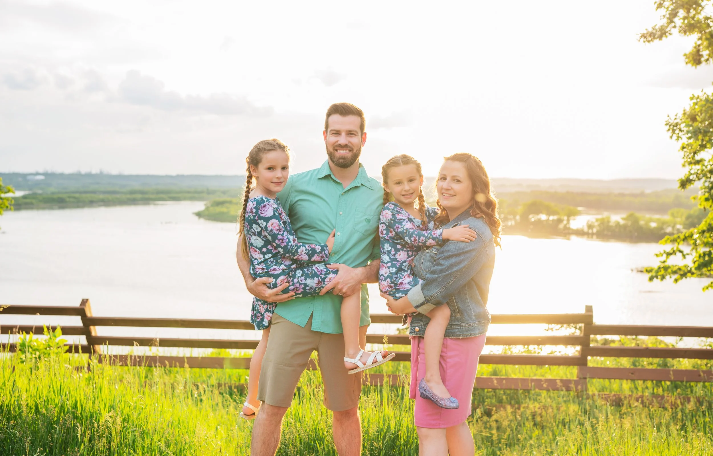 A family of four standing on a grassy hill near a lake during sunset, with two young girls in their parents' arms, all smiling at the camera.