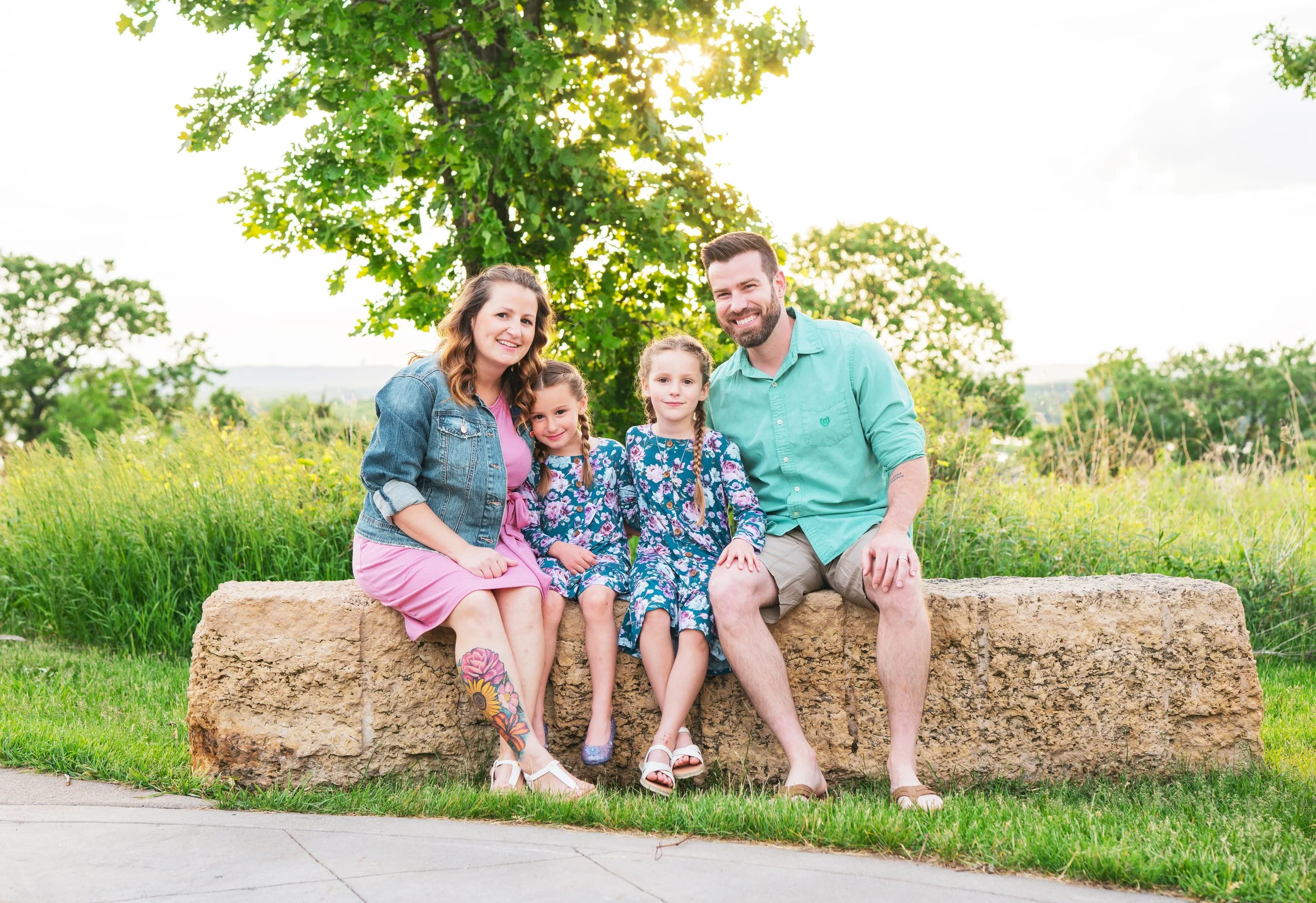 Family of four sitting on a stone bench outdoors in a park, with a green tree and grass in the background, smiling at the camera.