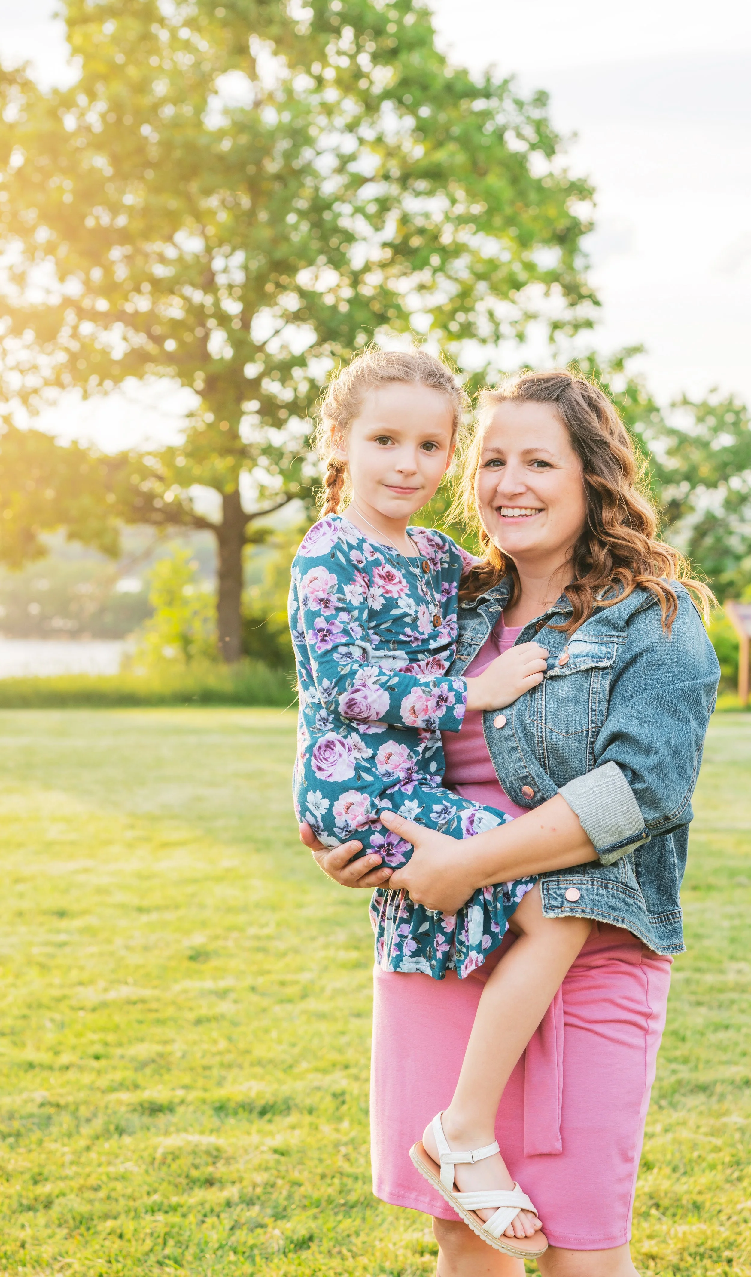 A smiling woman holding a young girl outdoors on a sunny day with green trees and grass in the background.