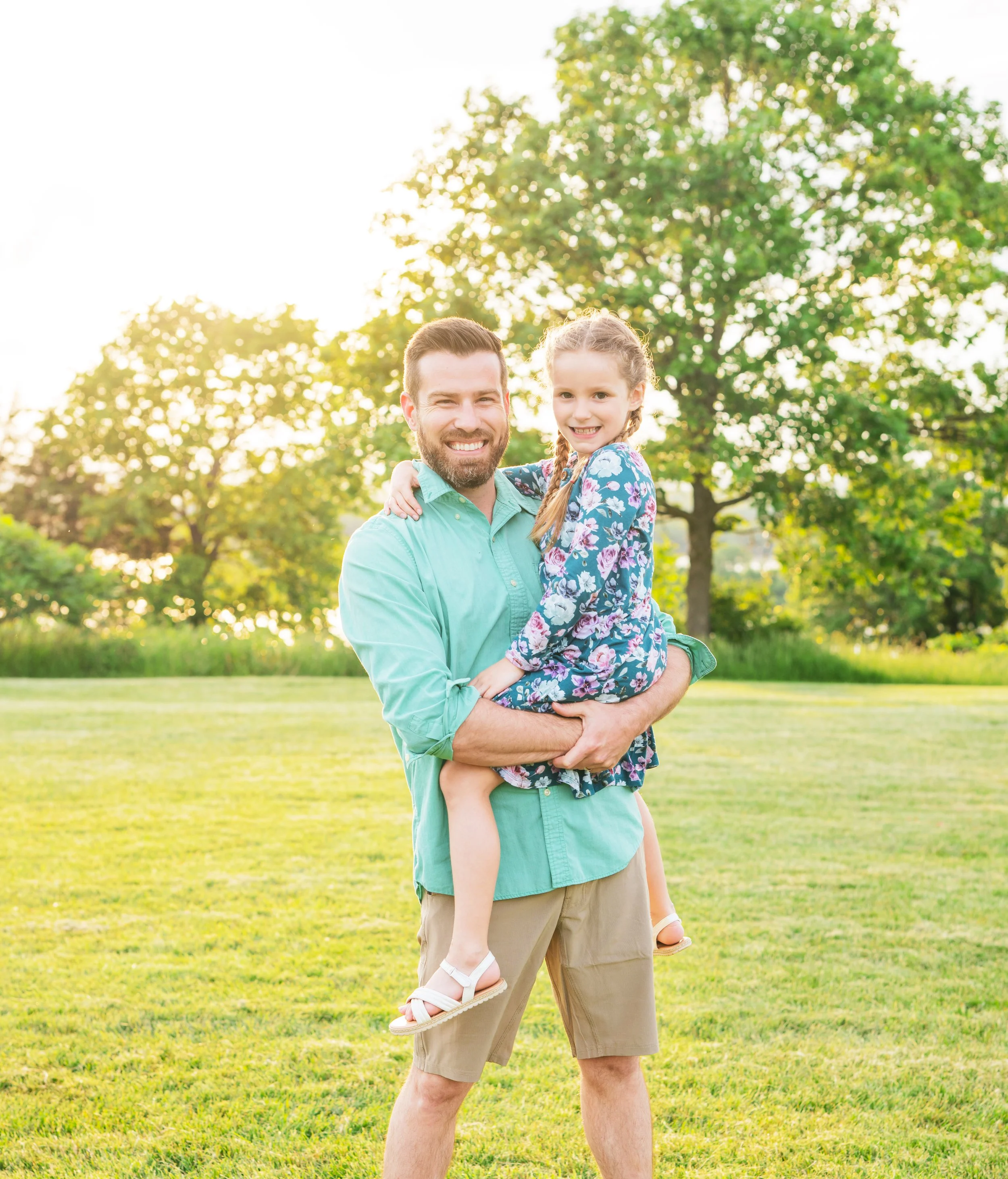 A man with a beard and a girl with braided hair smiling in a park during late afternoon, the man is holding the girl in his arms, and they are both looking at the camera. Trees and sunlight are in the background.