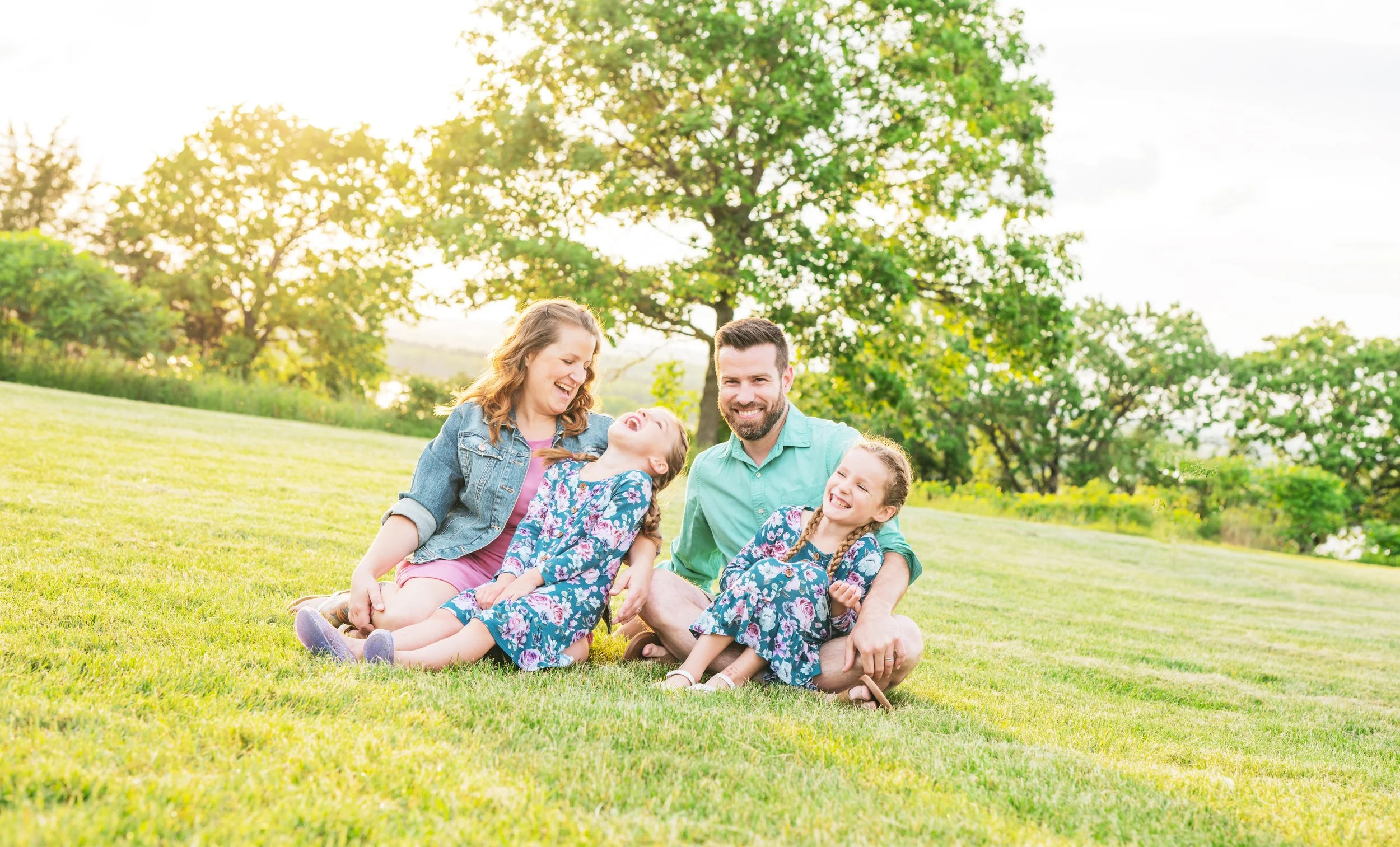 Family of four sitting and laughing on a grassy lawn in a park with trees, enjoying a sunny day.