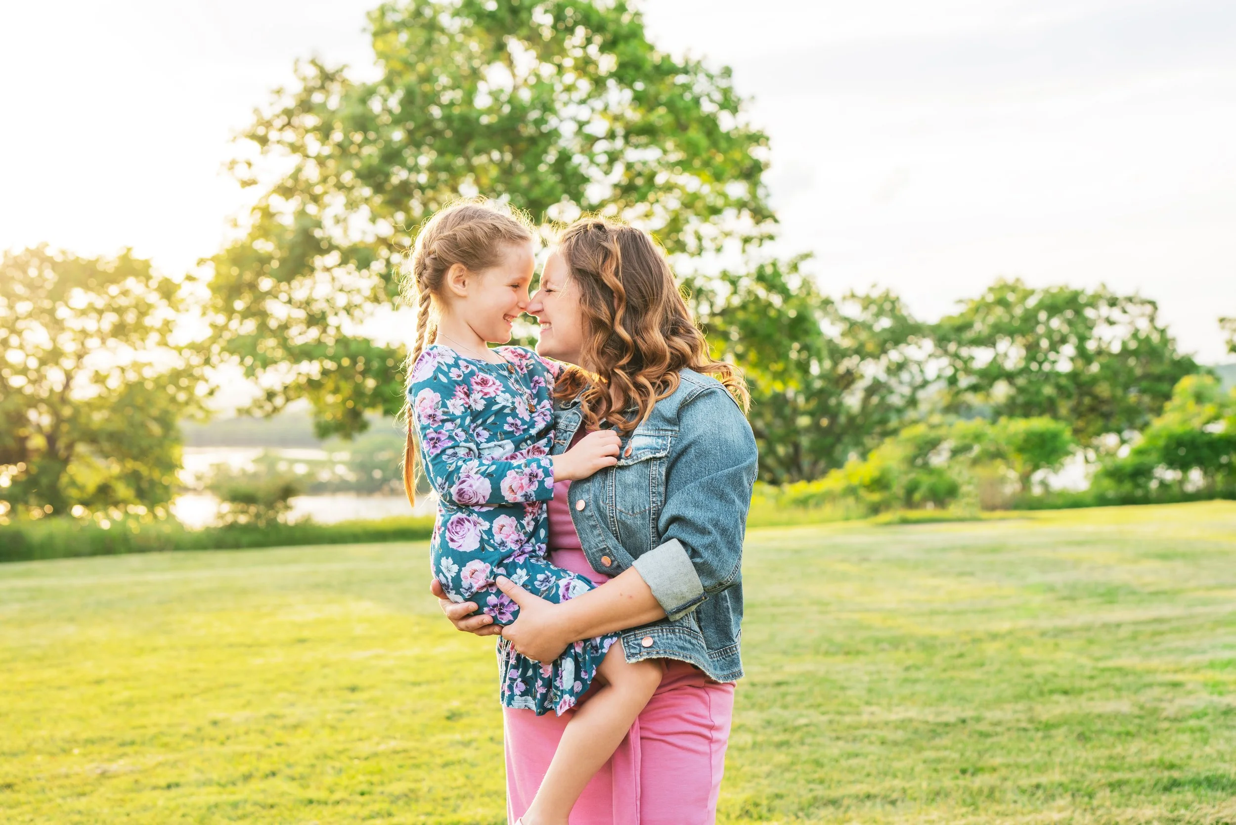A woman holding a young girl in a park at sunset, both smiling with noses touching, surrounded by green trees and grass.