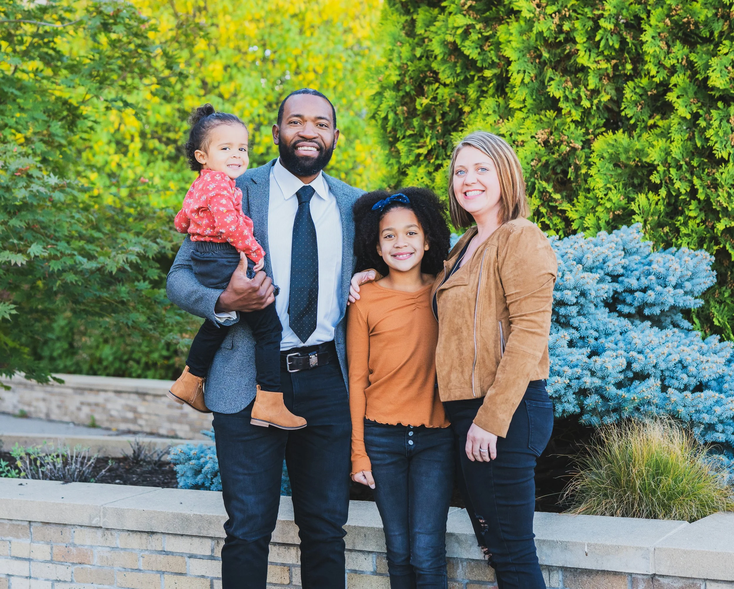 A family of four standing outdoors in front of green trees and bushes. The father is holding a young girl, and a woman is standing beside a teenage girl. Everyone is smiling and wearing casual autumn clothing.